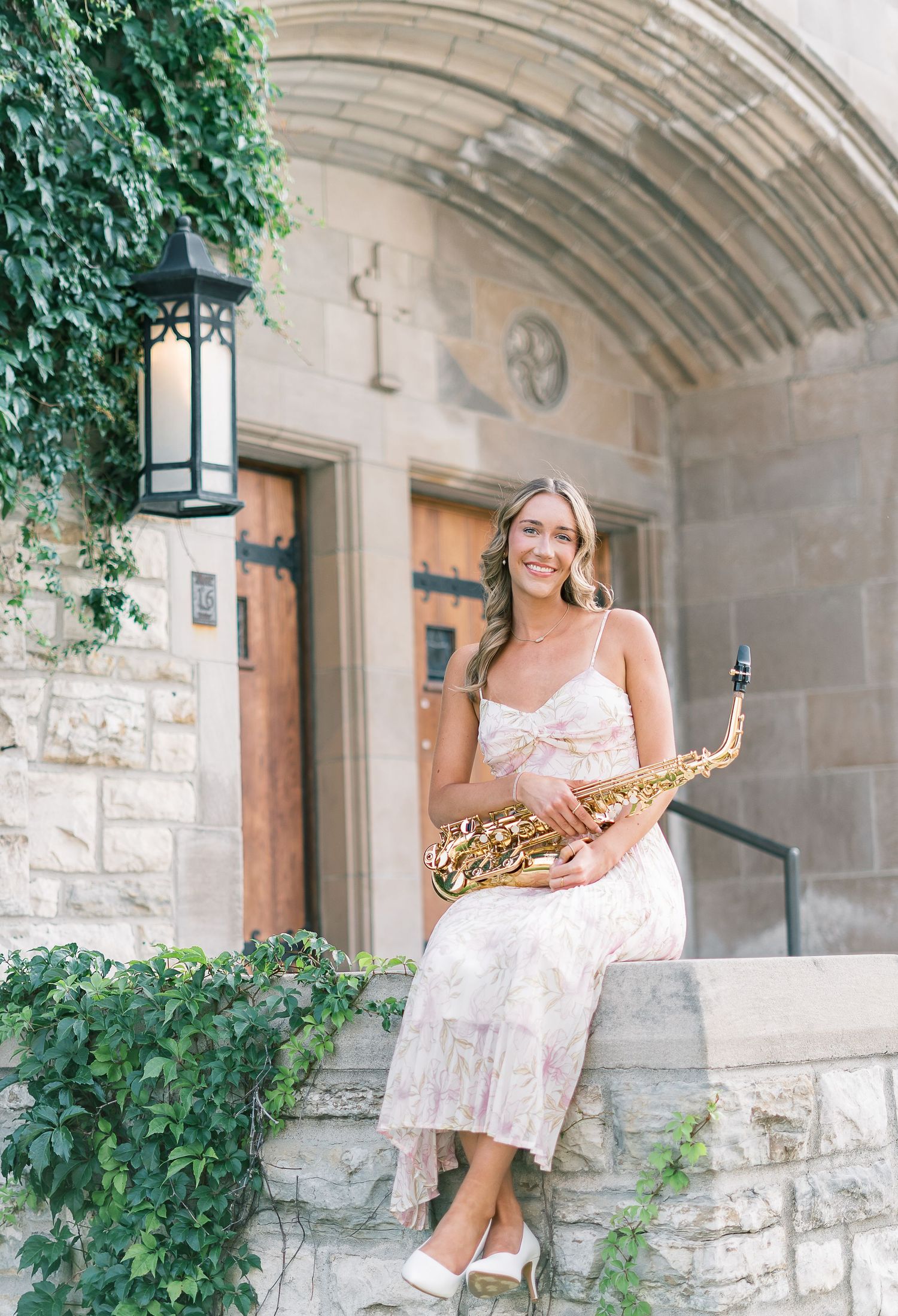 Musician in a white dress sits on stone steps with a saxophone in a garden archway setting.
