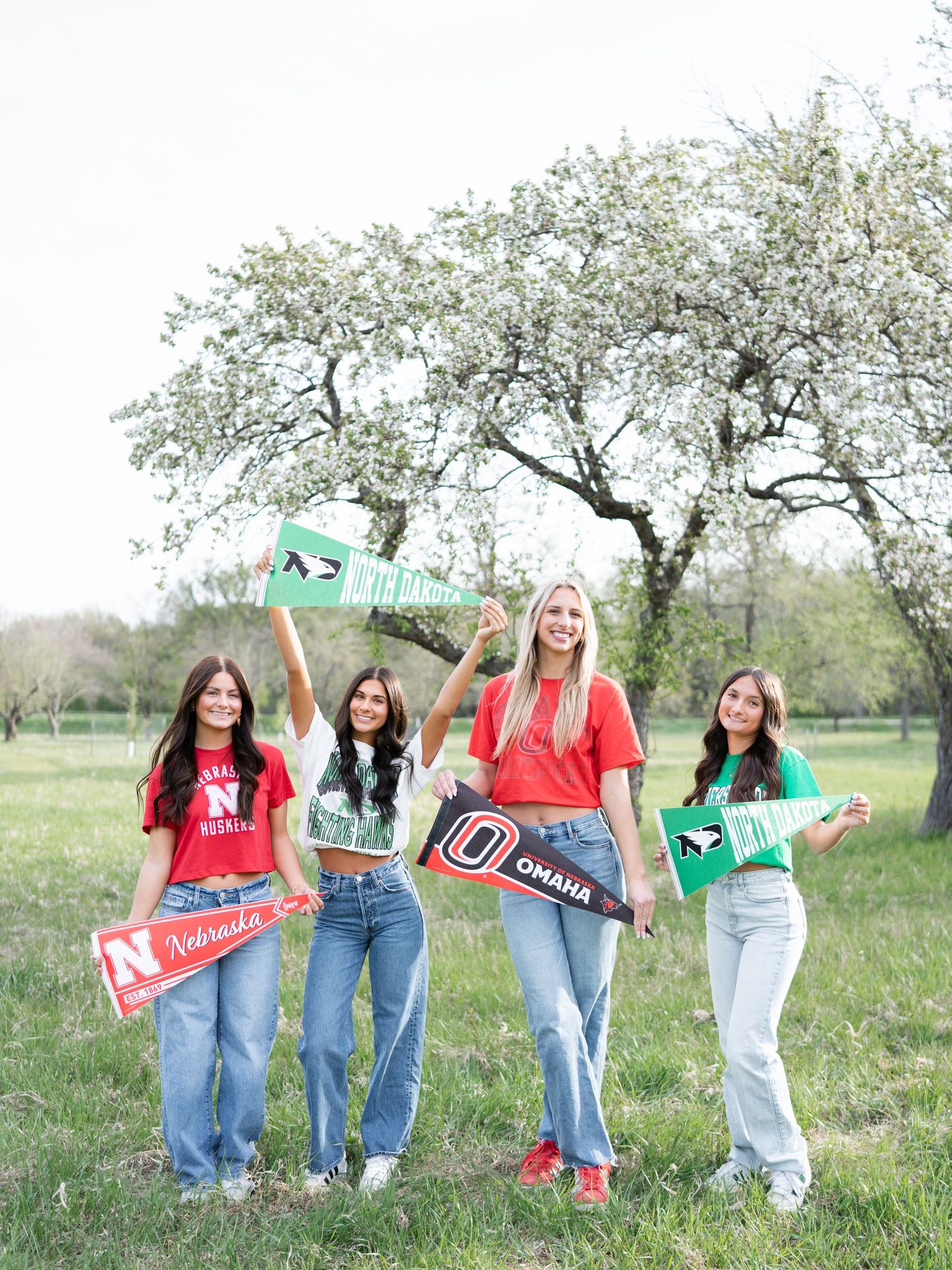 Four college girls holding university pennants and signs outdoors near a blooming tree in a green field.