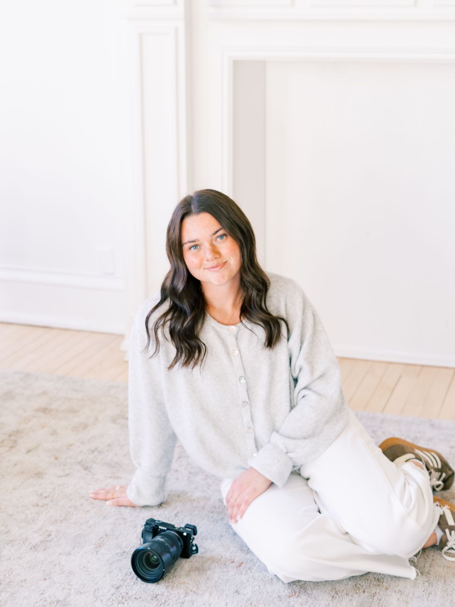 A woman with dark hair sits on a white floor smiling, holding a DSLR camera in a bright minimalist studio.