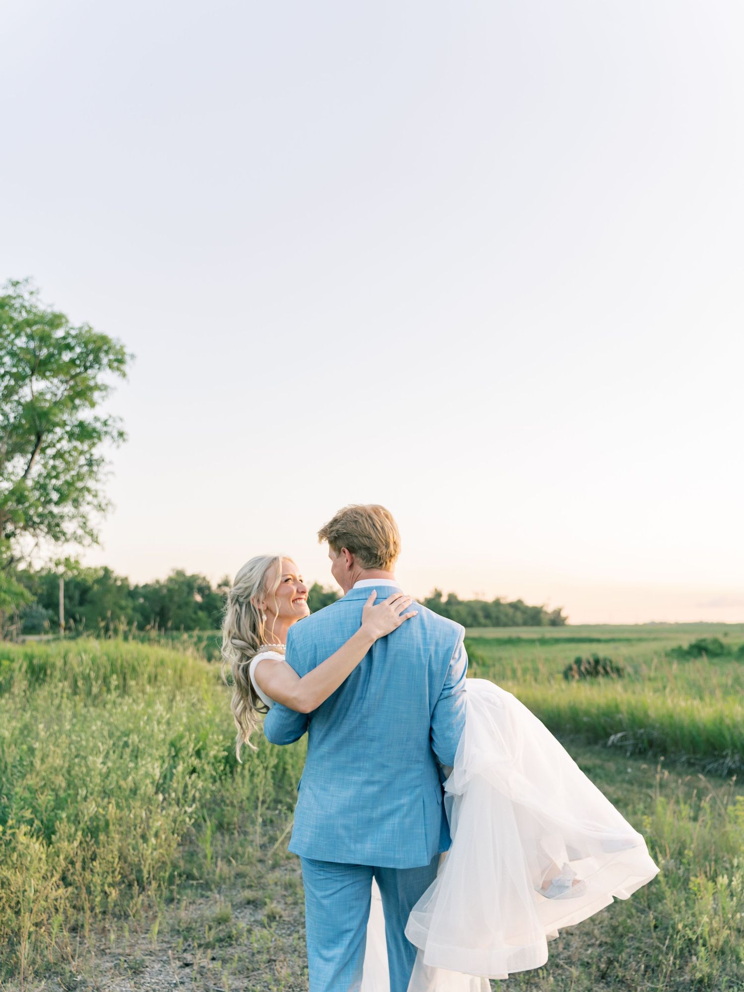 Groom in blue suit lifts bride in white dress in a green field at sunset.