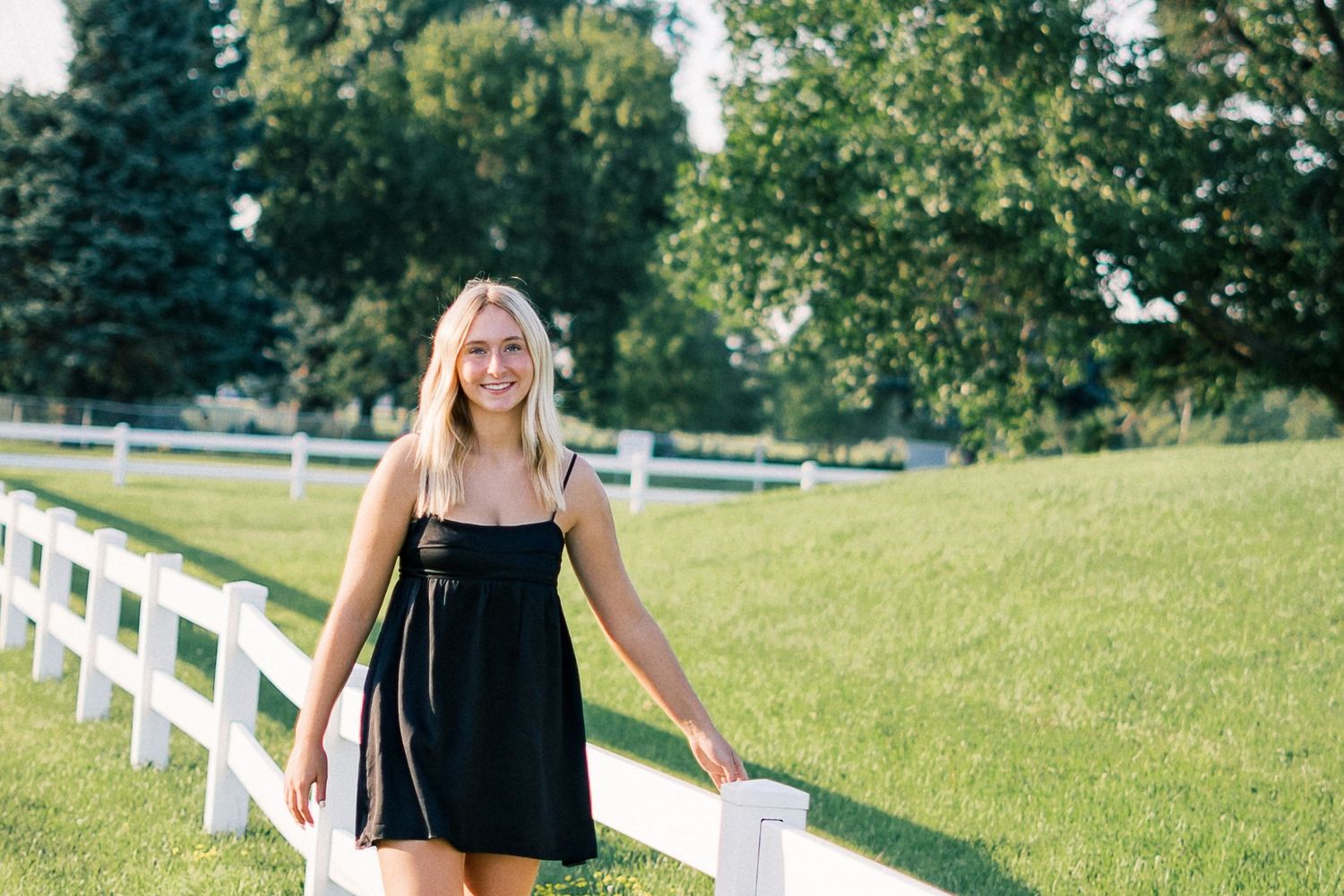Woman in black dress stands near white fence on grassy hill during sunny summer day.