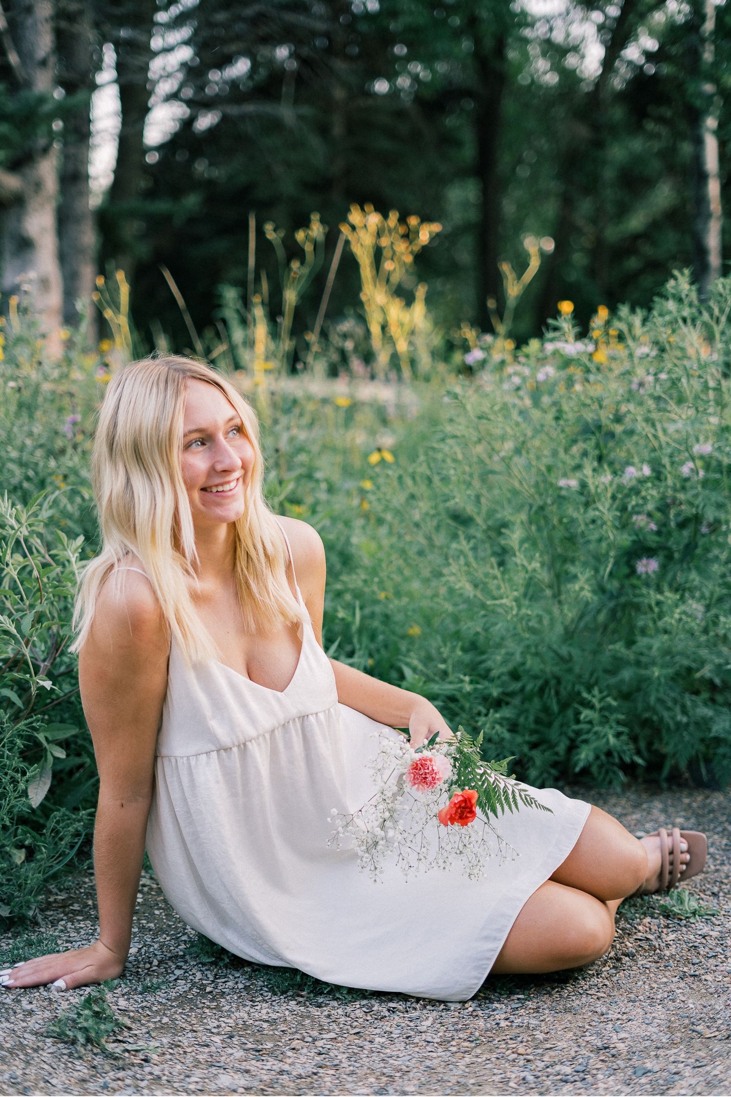 Woman in white summer dress relaxes on ground among wildflowers in outdoor nature setting.