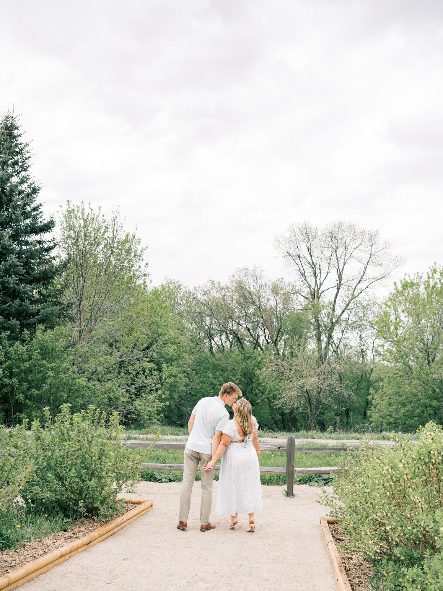 Couple sharing a romantic kiss on a garden path surrounded by lush green trees and shrubs on a cloudy day.
