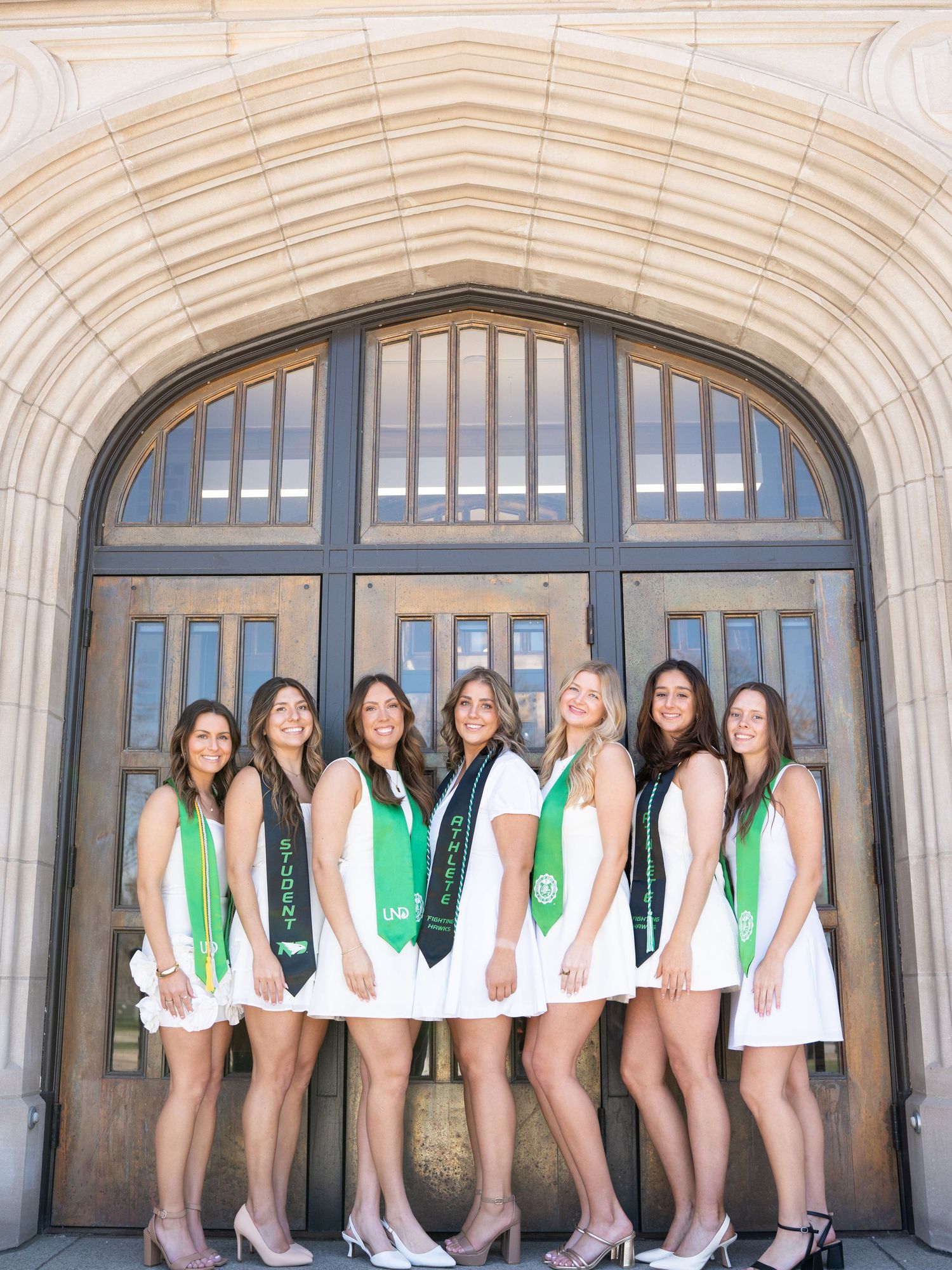 Seven female graduates in white dresses and green sashes pose together under an arched stone doorway.