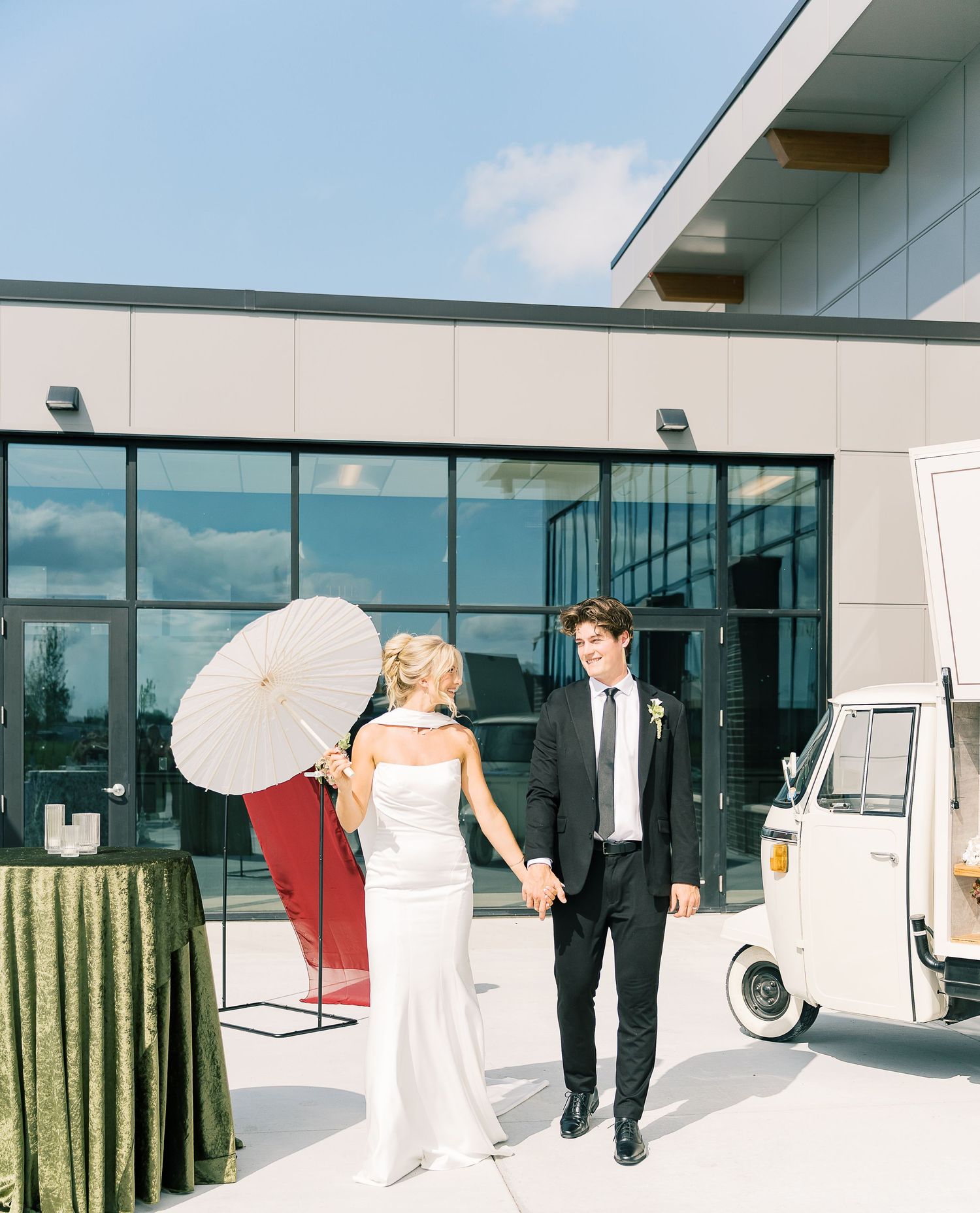 A couple in wedding attire walking past a modern building exterior with floor-to-ceiling windows.