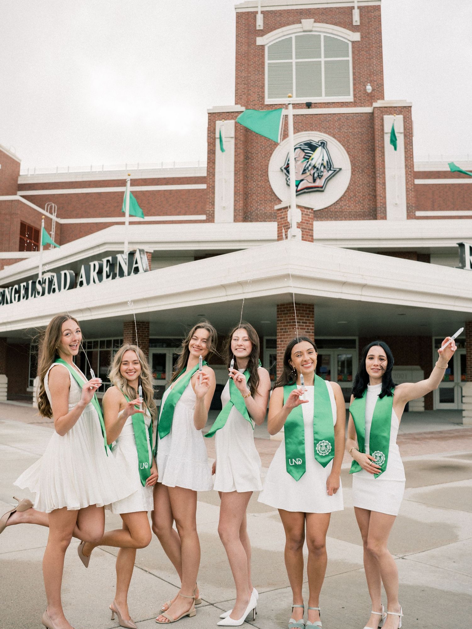 Six women in white dresses and green graduation stoles celebrate outside Englestad Arena on graduation day.