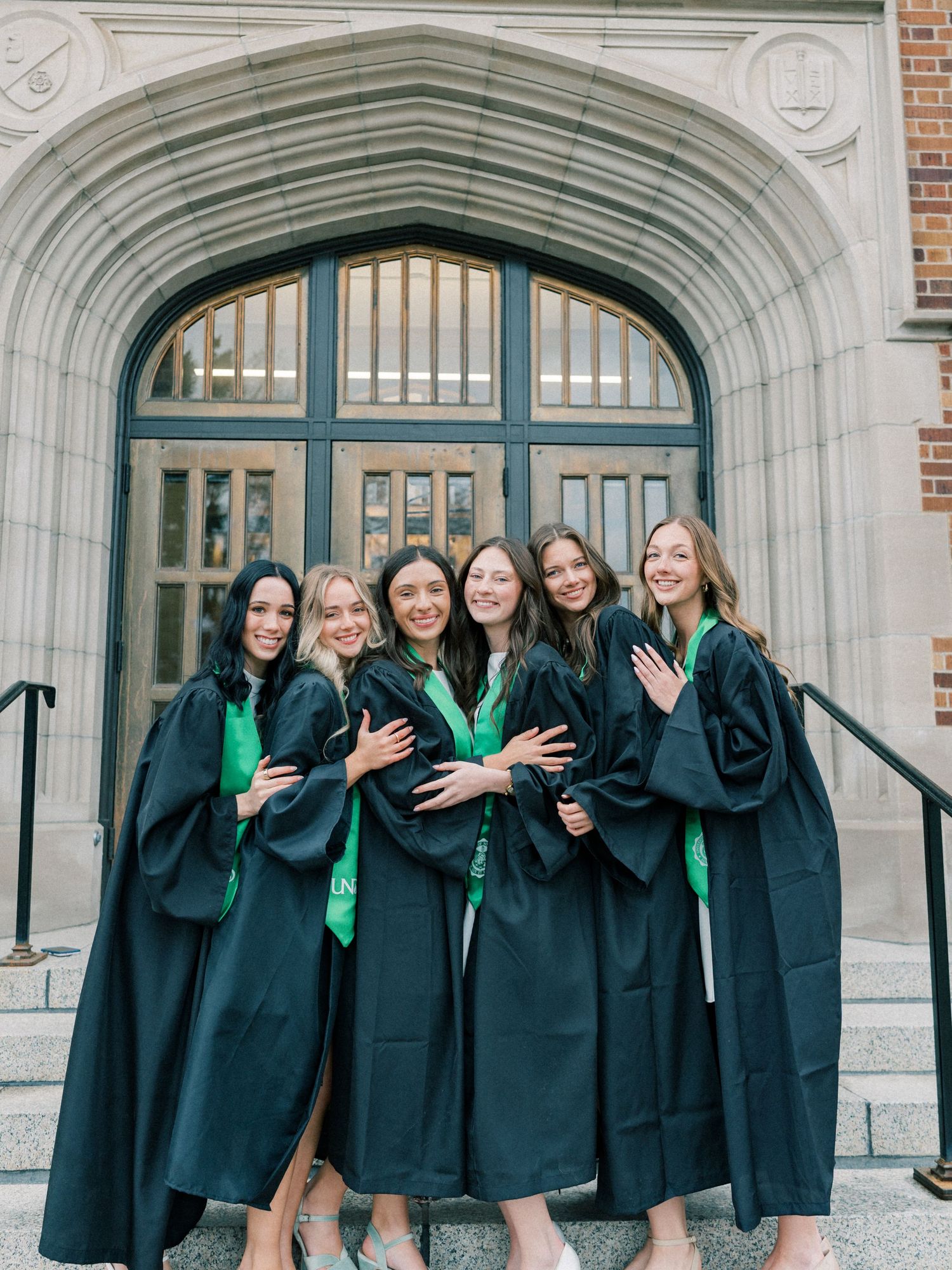 Six women in black graduation gowns with green stoles pose together on steps outside a stone building.