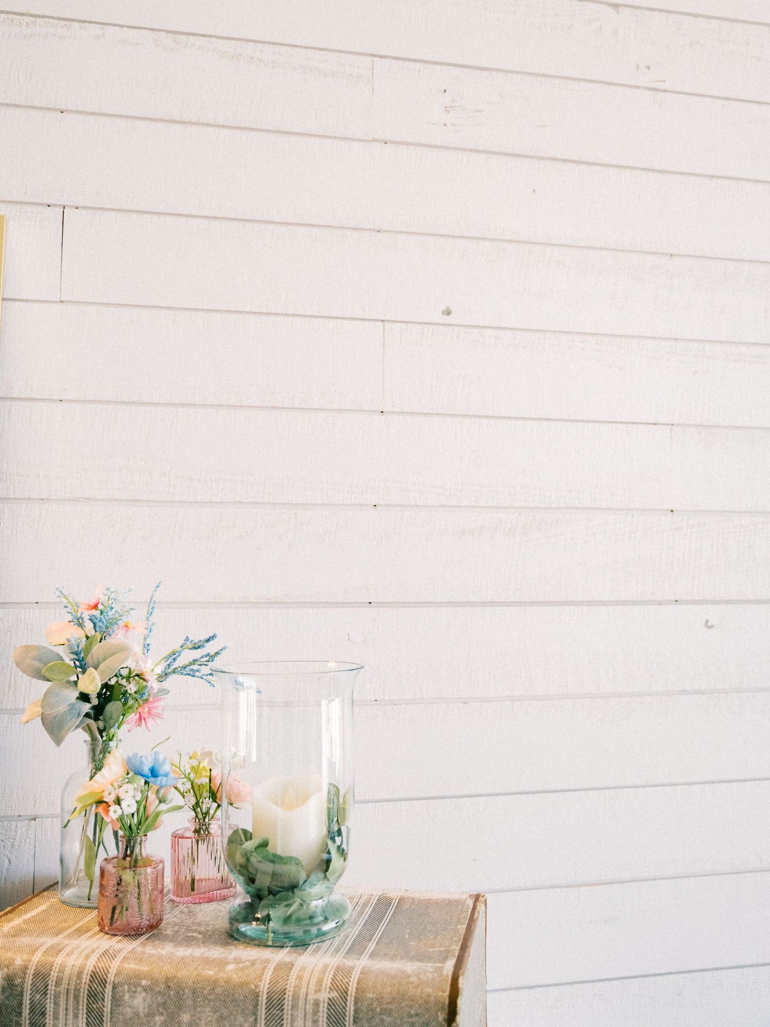 Colorful spring flowers in glass vases on a burlap table runner against a white shiplap wall.