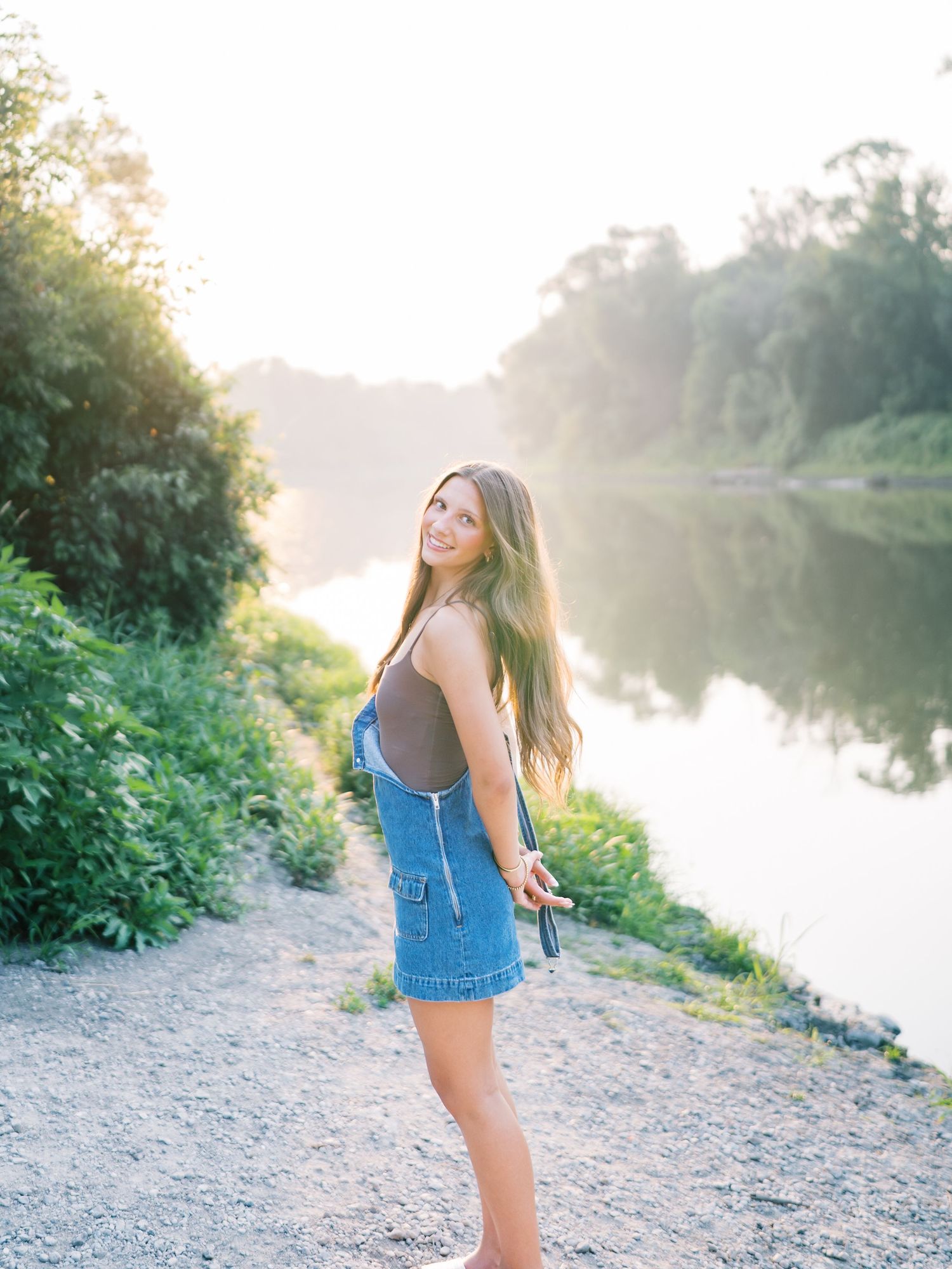 Young woman in denim overalls smiling by a serene river at golden hour, surrounded by lush greenery.