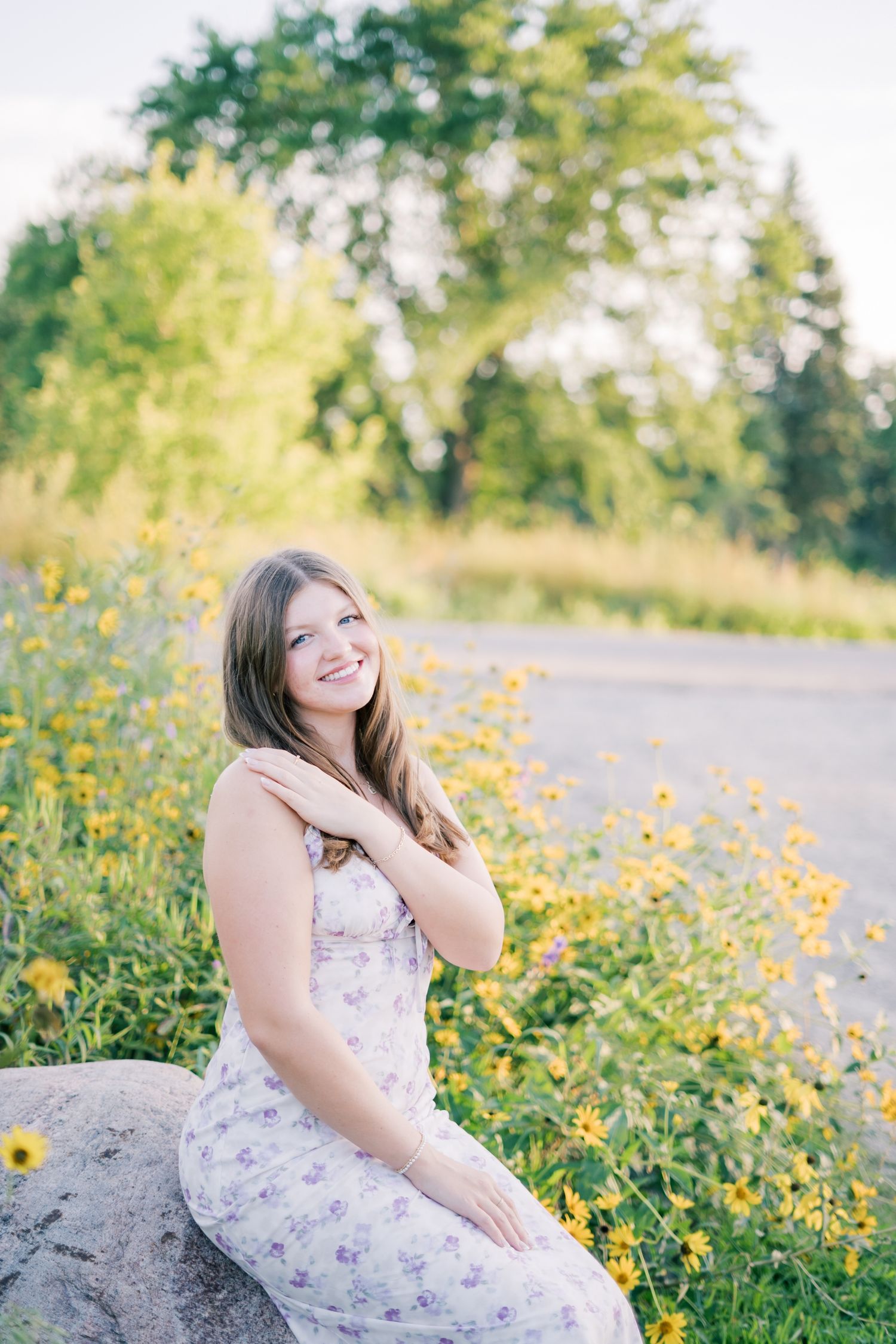 A woman in a floral dress sits on a large rock surrounded by blooming yellow wildflowers in a summer field.