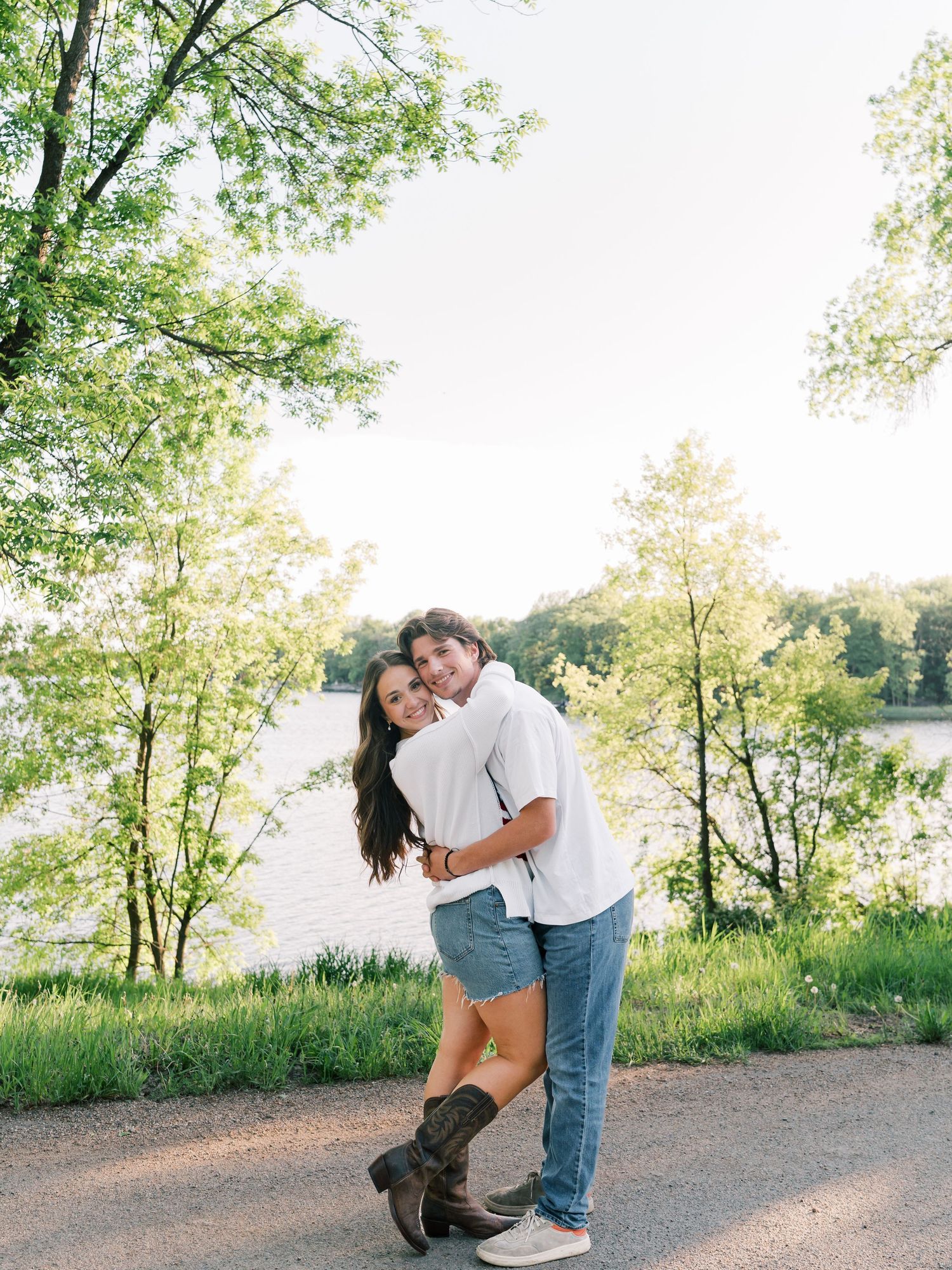 Couple embracing and smiling outdoors near a lake, surrounded by lush green trees during golden hour.