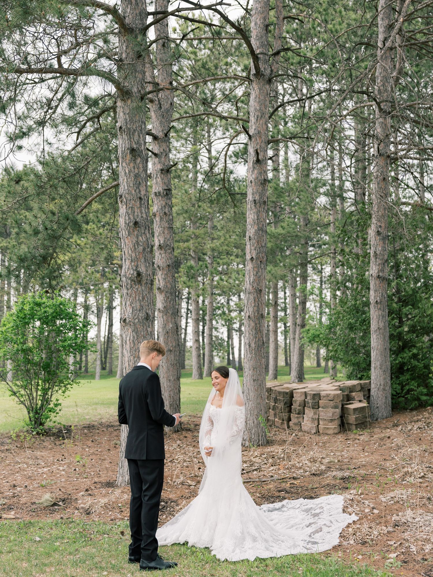 Bride and groom share a first look moment among tall pine trees, bride wearing a lace mermaid gown with long train.