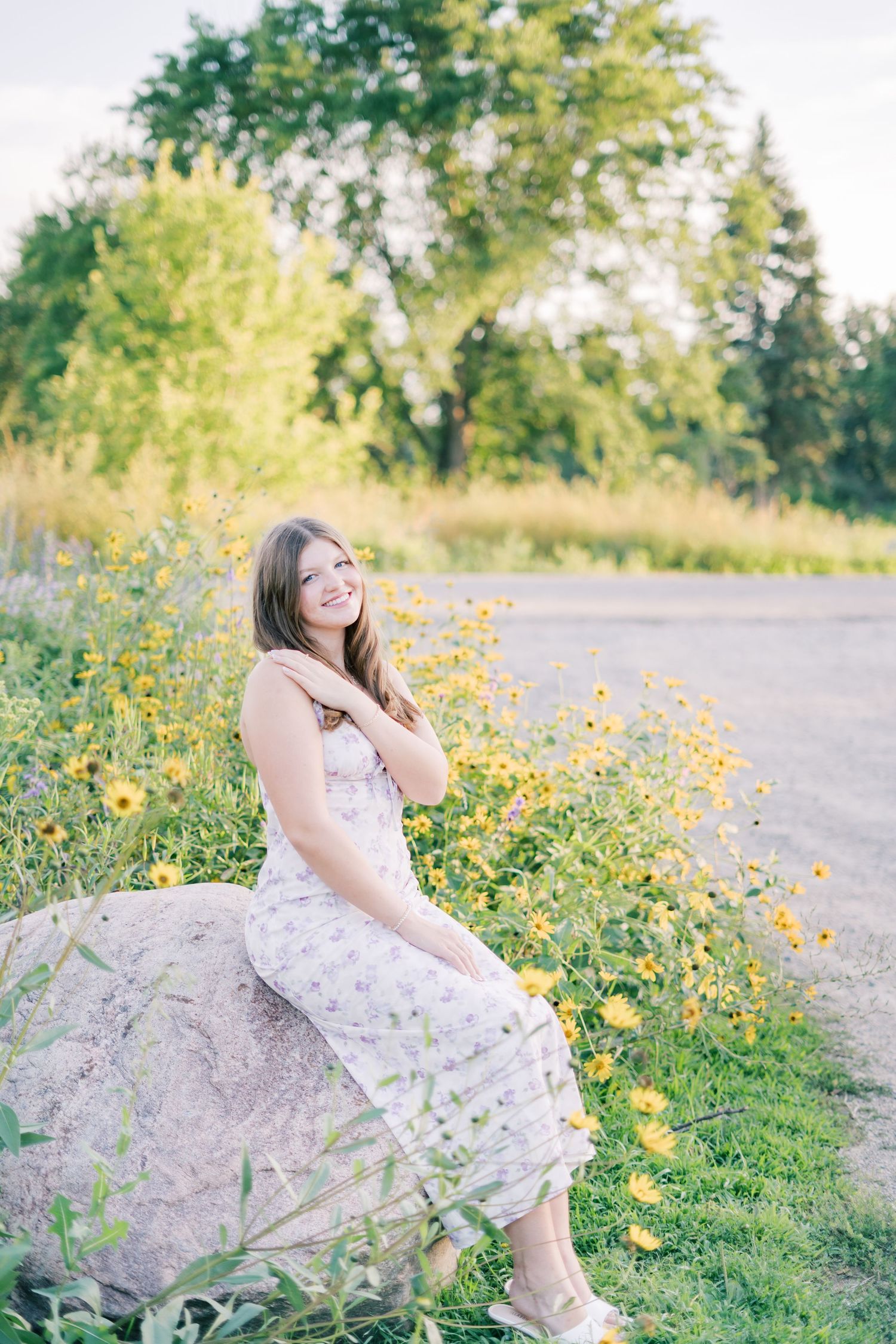 A person in a white floral dress sits on a large rock near a field of yellow wildflowers on a sunny summer day.