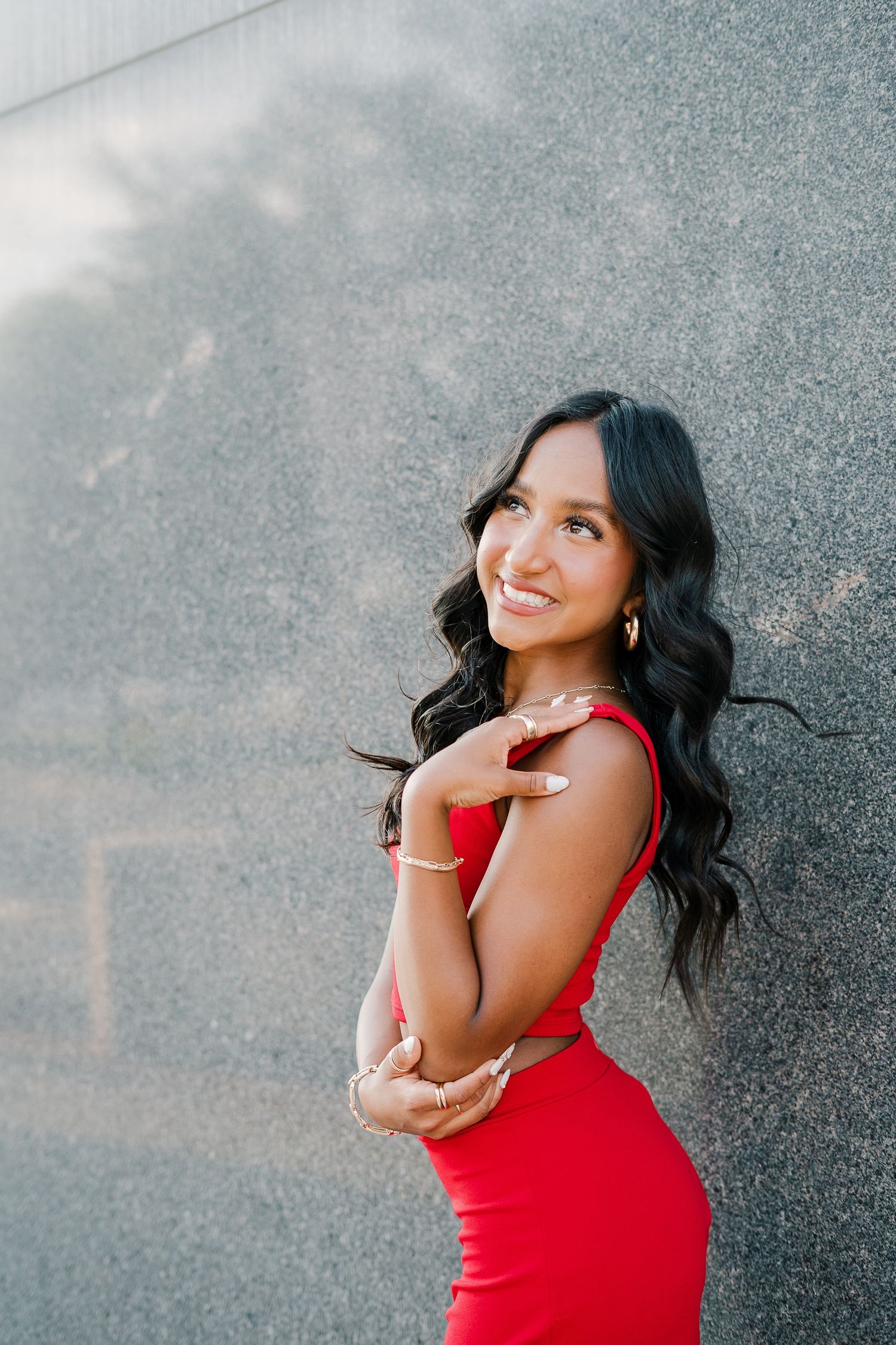 Woman in red sleeveless dress poses against textured gray wall with joyful expression in outdoor portrait series.