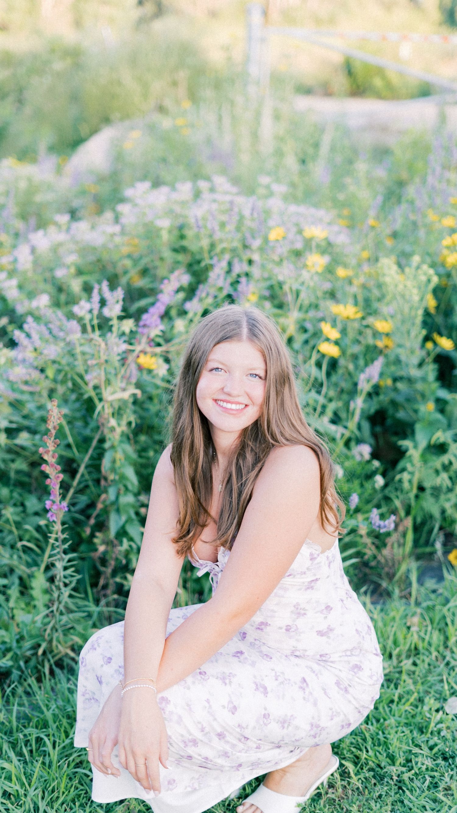 A person in a white floral dress poses joyfully among wildflowers in a sunny meadow.