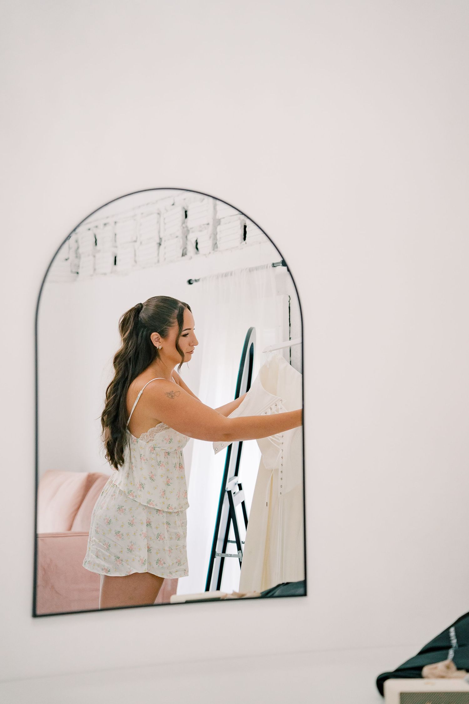 A series of arched mirrors on a white wall reflect someone in a light-colored dress reaching to adjust them.