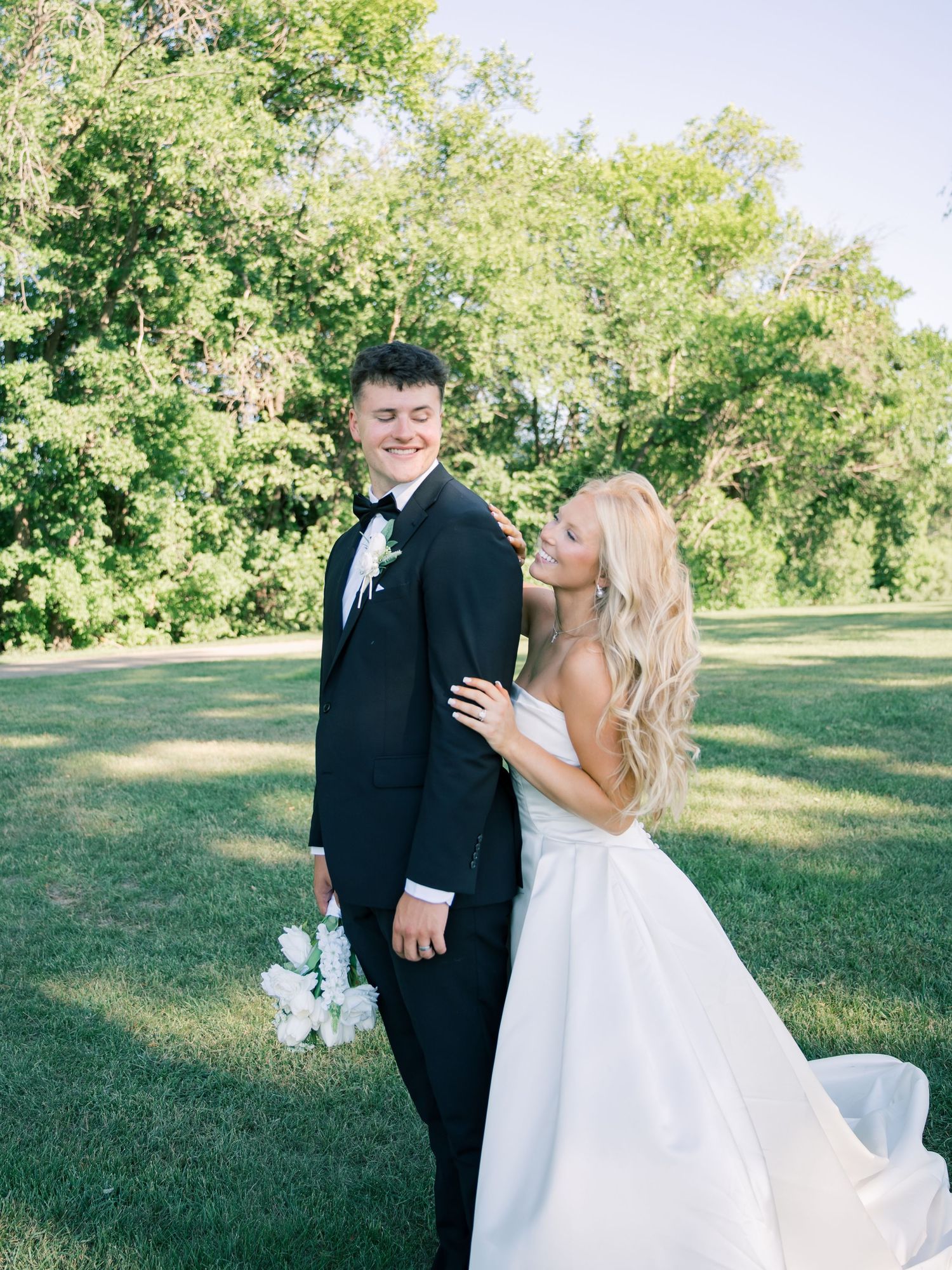 A bride in a white ballgown and groom in a black tuxedo pose together on a lush green lawn surrounded by trees.