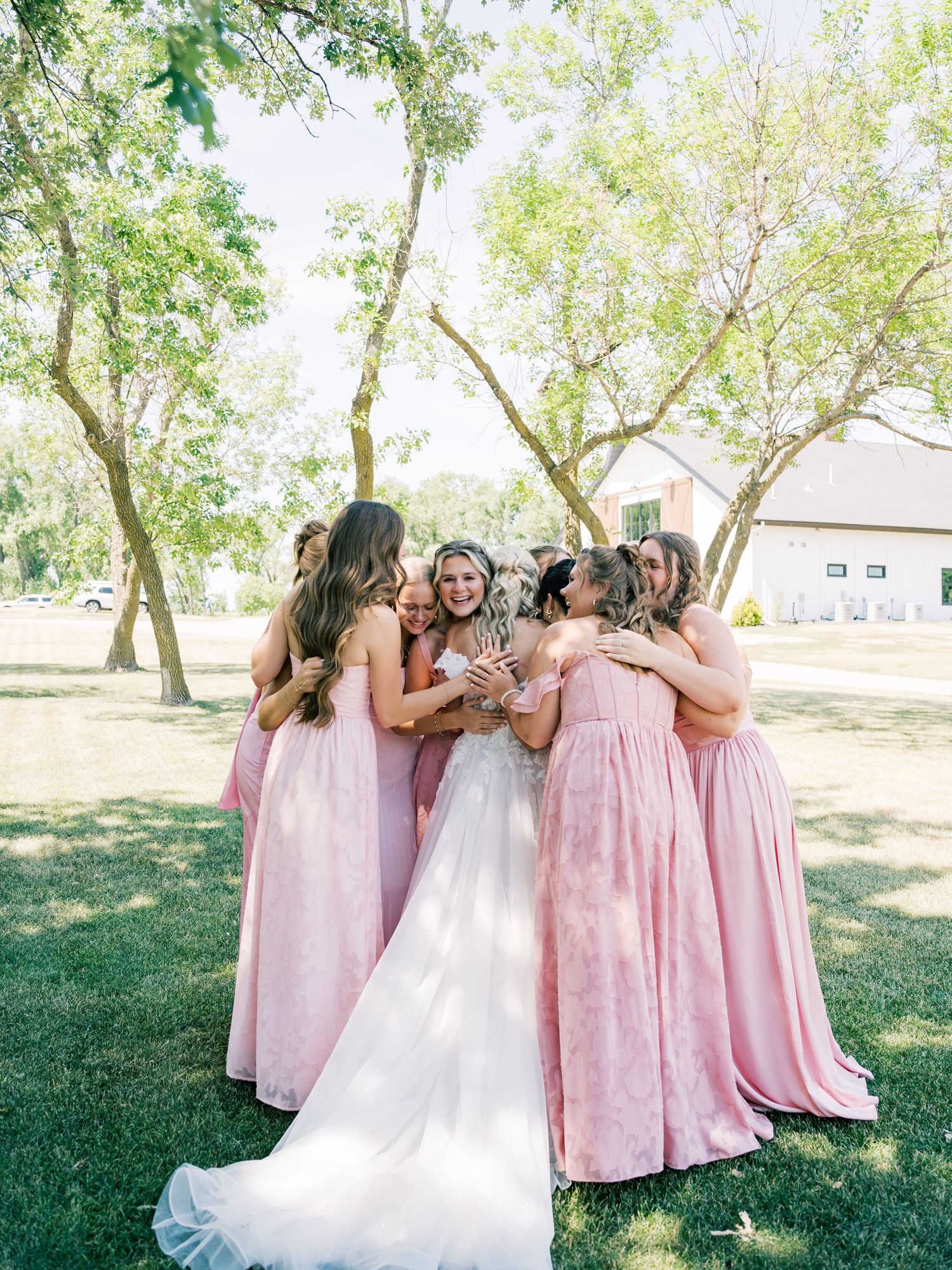 Bride in white gown hugged by bridesmaids in pink dresses on green lawn with trees and white barn.
