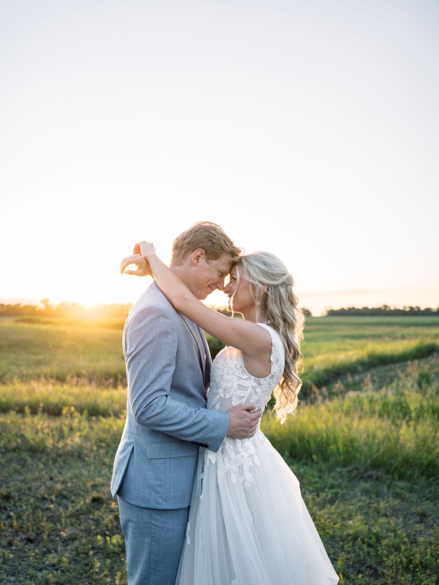 Bride and groom embrace at golden sunset in green field, wearing lace wedding gown and light gray suit.