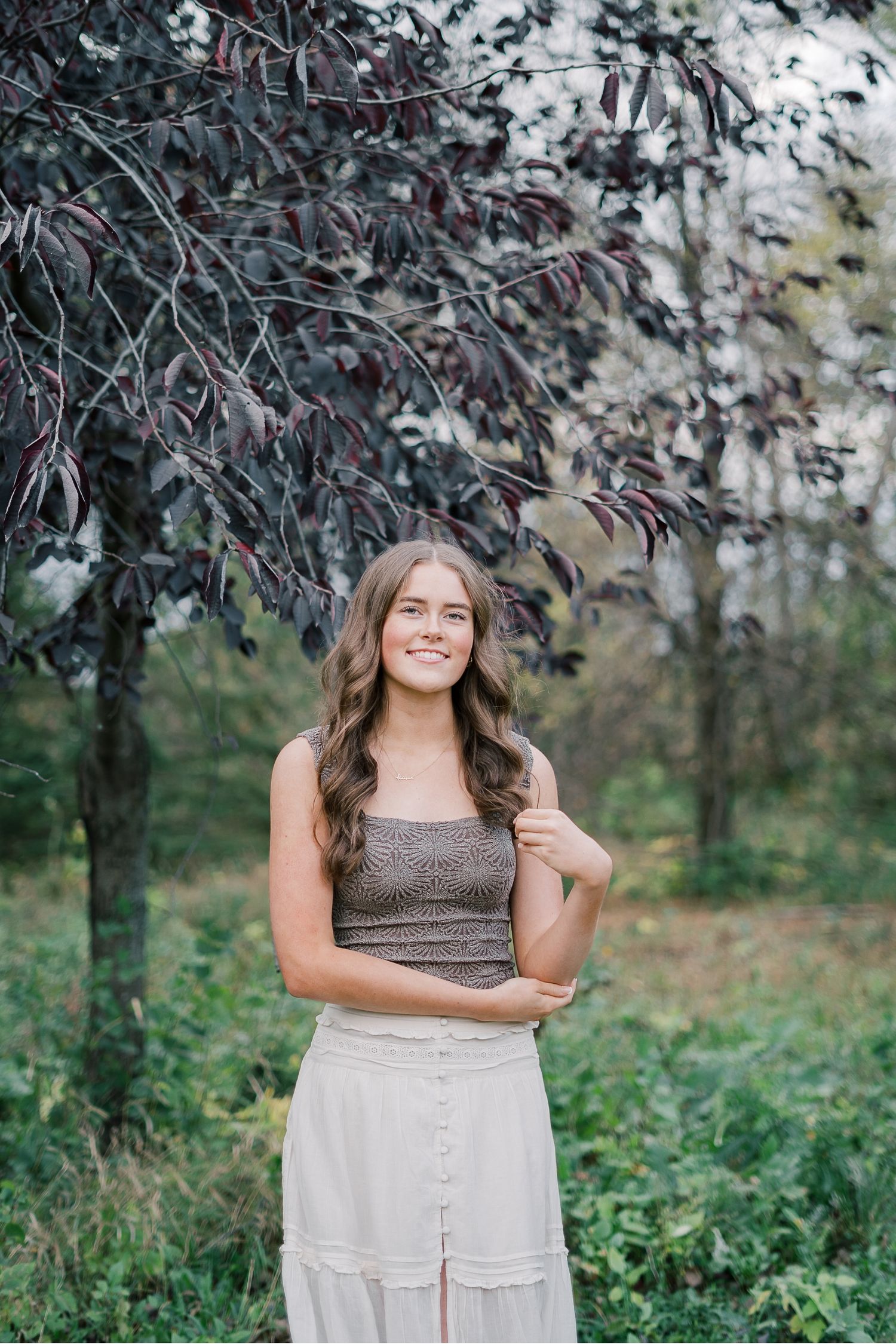 A person wearing a brown crop top and white button-up skirt poses outdoors under flowering trees in a natural setting.