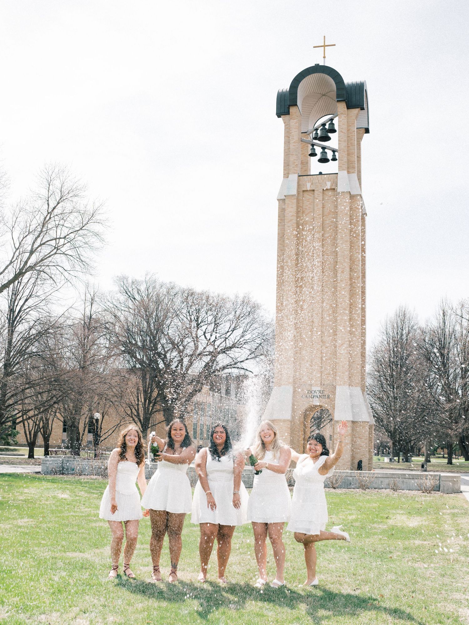 Five women in white dresses celebrate outdoors near a tall brick bell tower with a cross on top.