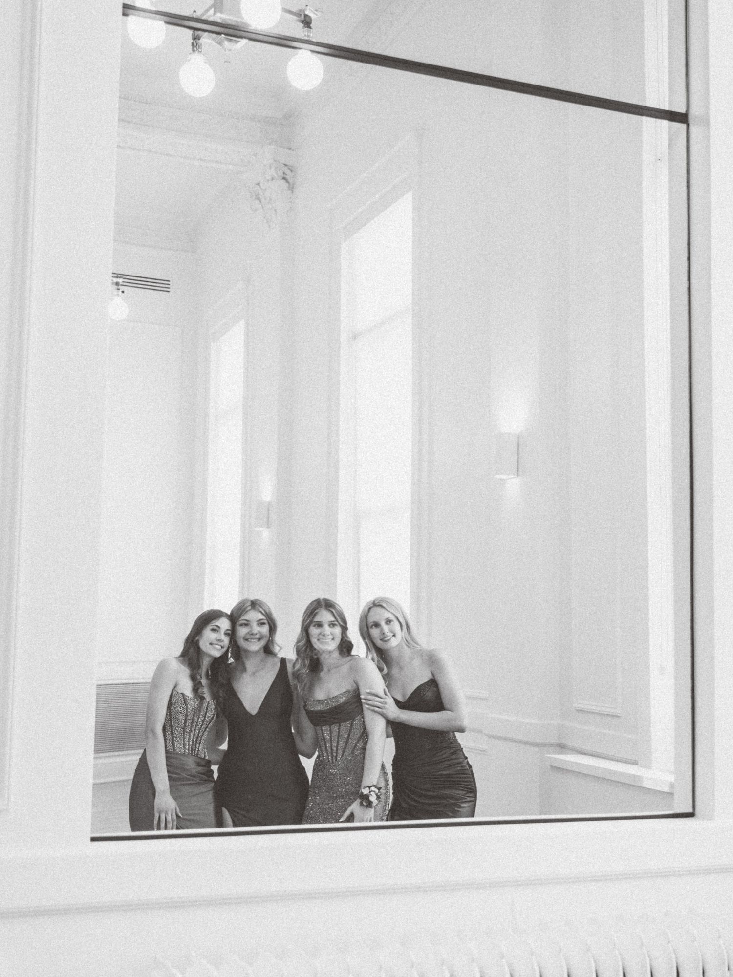 Four women in elegant black dresses pose together smiling in a mirror reflection at a formal event.