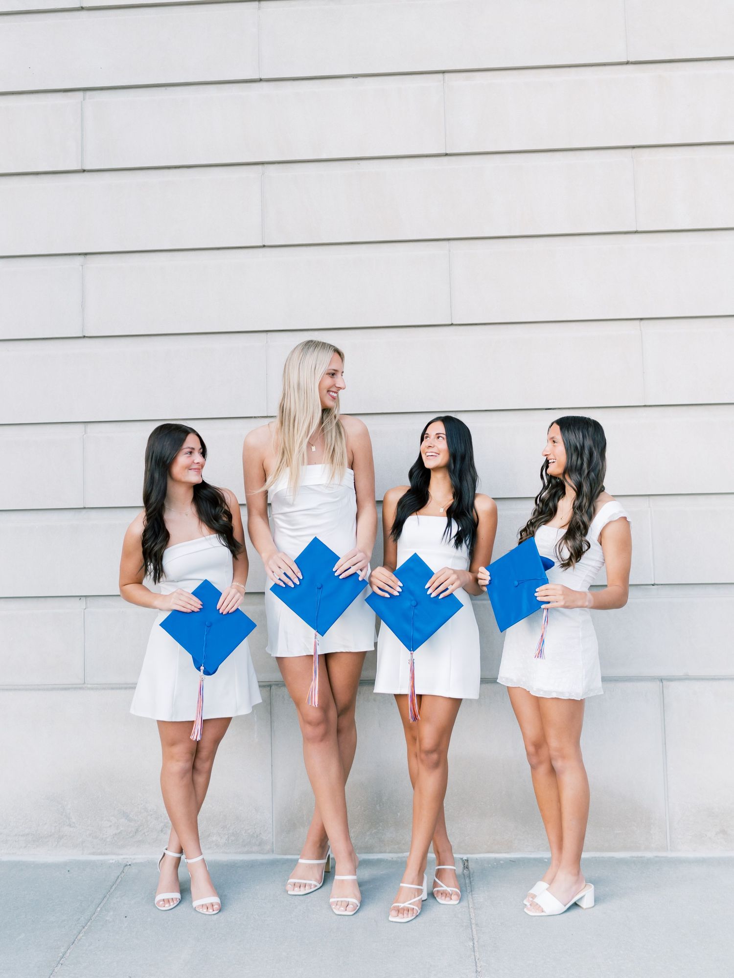 Four women in white dresses holding blue graduation caps pose together against a stone wall.