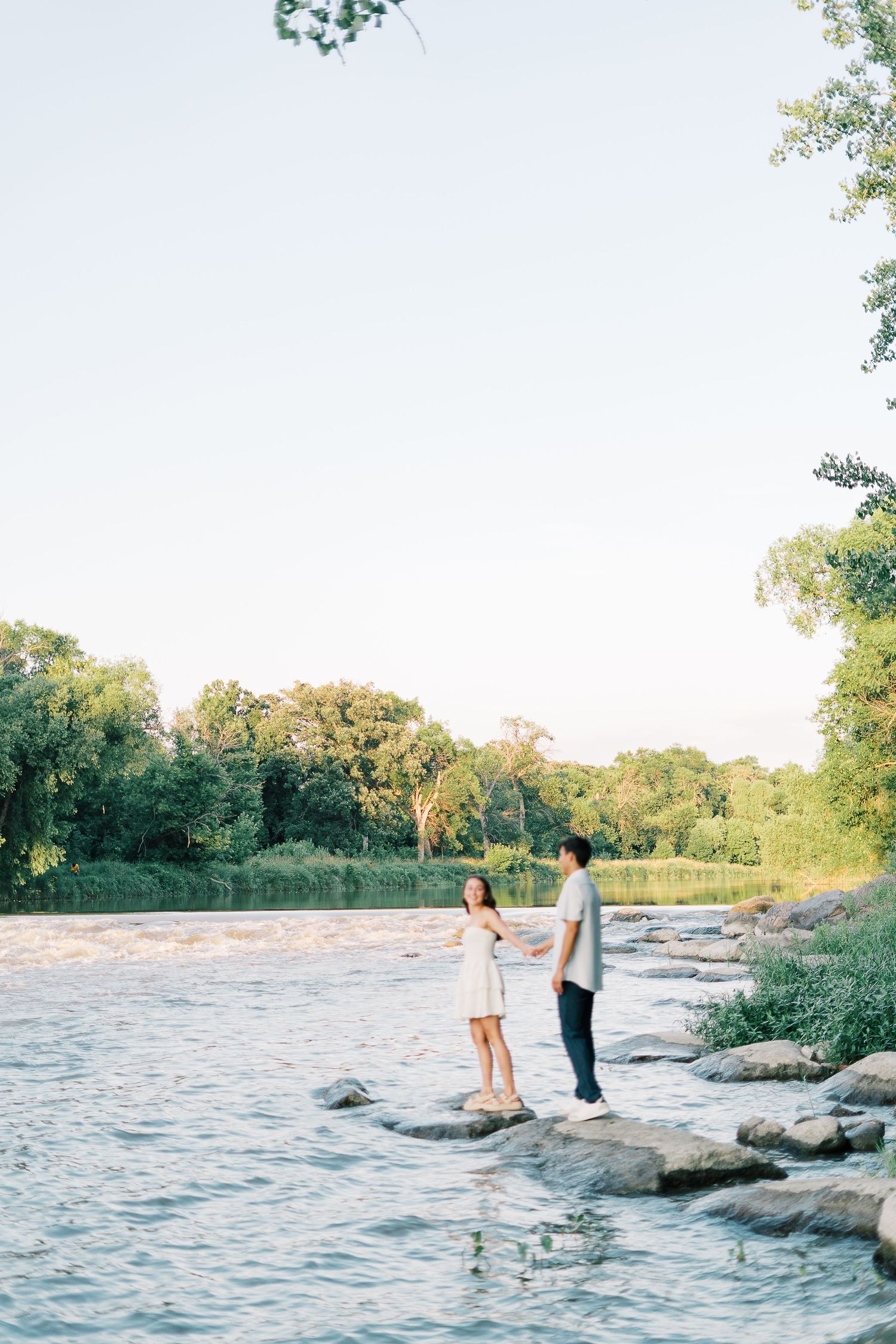 Couple walking along a rocky shoreline at sunset with trees in the background.