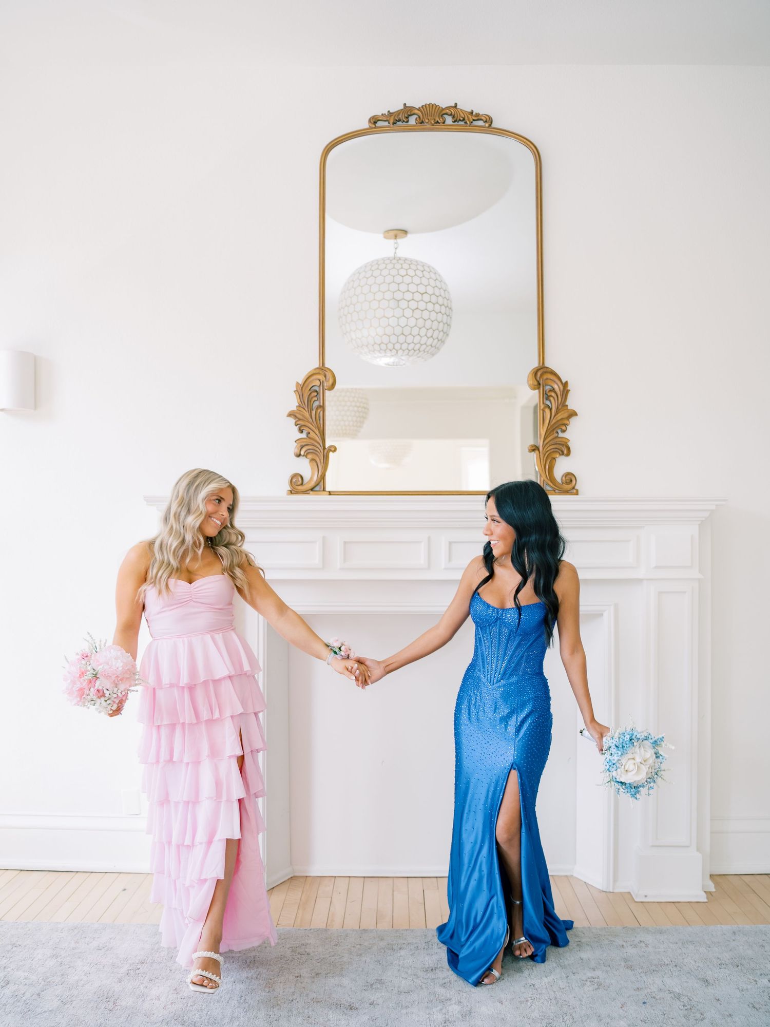 Two women in pink and blue gowns hold hands before a gold ornate mirror above a white fireplace.