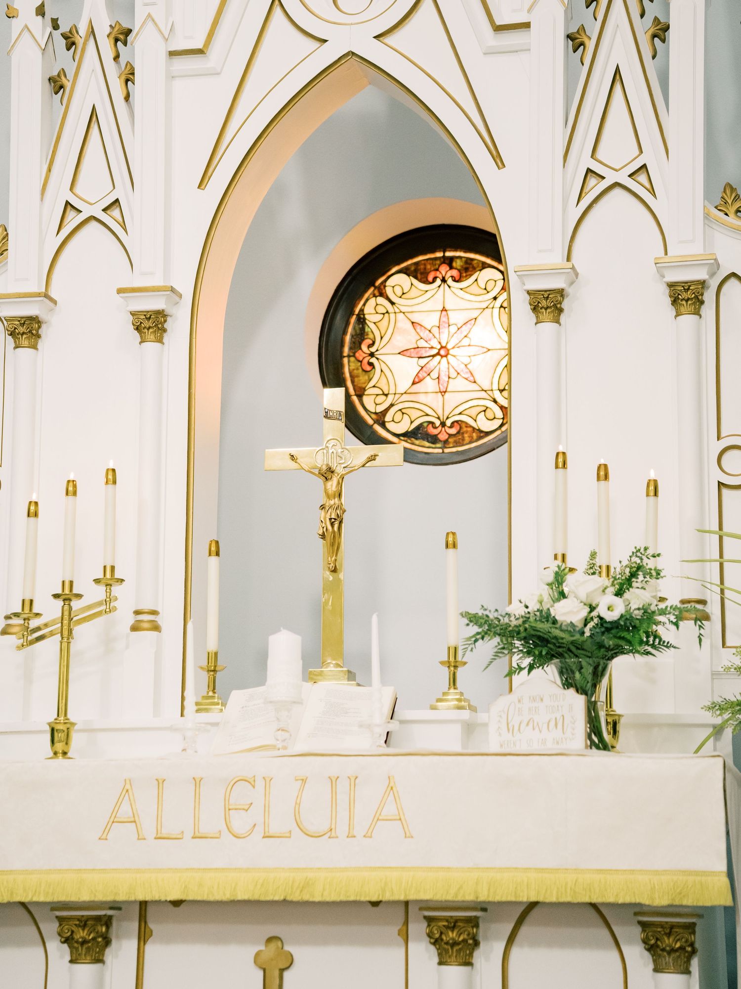 Elegant church altar with gold accents, crucifix, candles, white floral arrangement, and ALLELUIA inscription in a white Gothic setting.