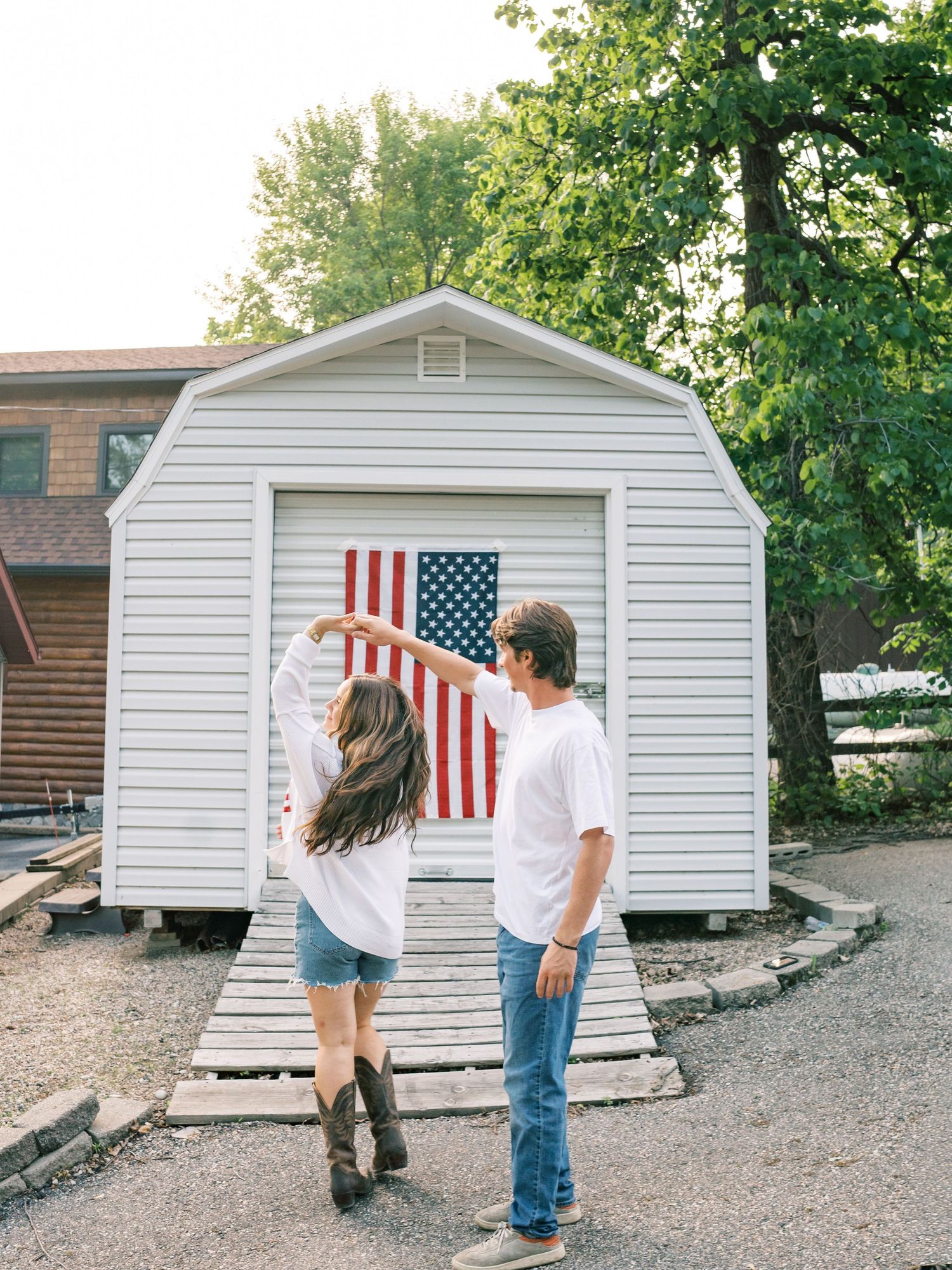 Couple dancing in front of a white shed with an American flag, woman in cowboy boots and denim shorts, man in white tee.