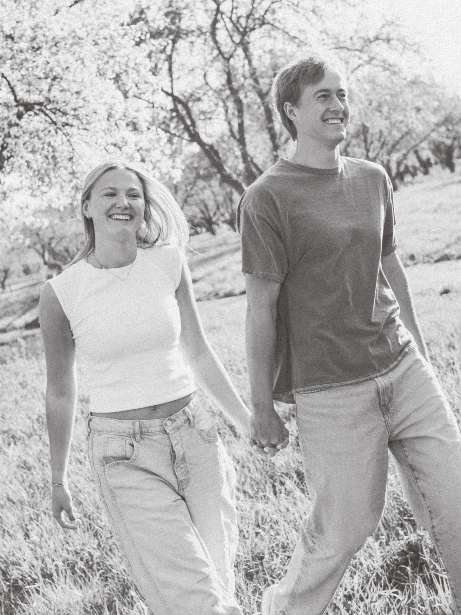 A smiling couple holds hands while walking outdoors in a field with trees in the background, black and white photo.