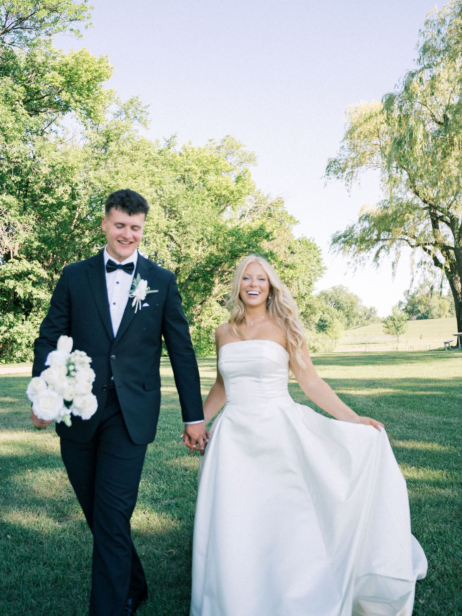 A joyful bride in a white strapless gown and groom in black tuxedo walk hand in hand on lush green grass.
