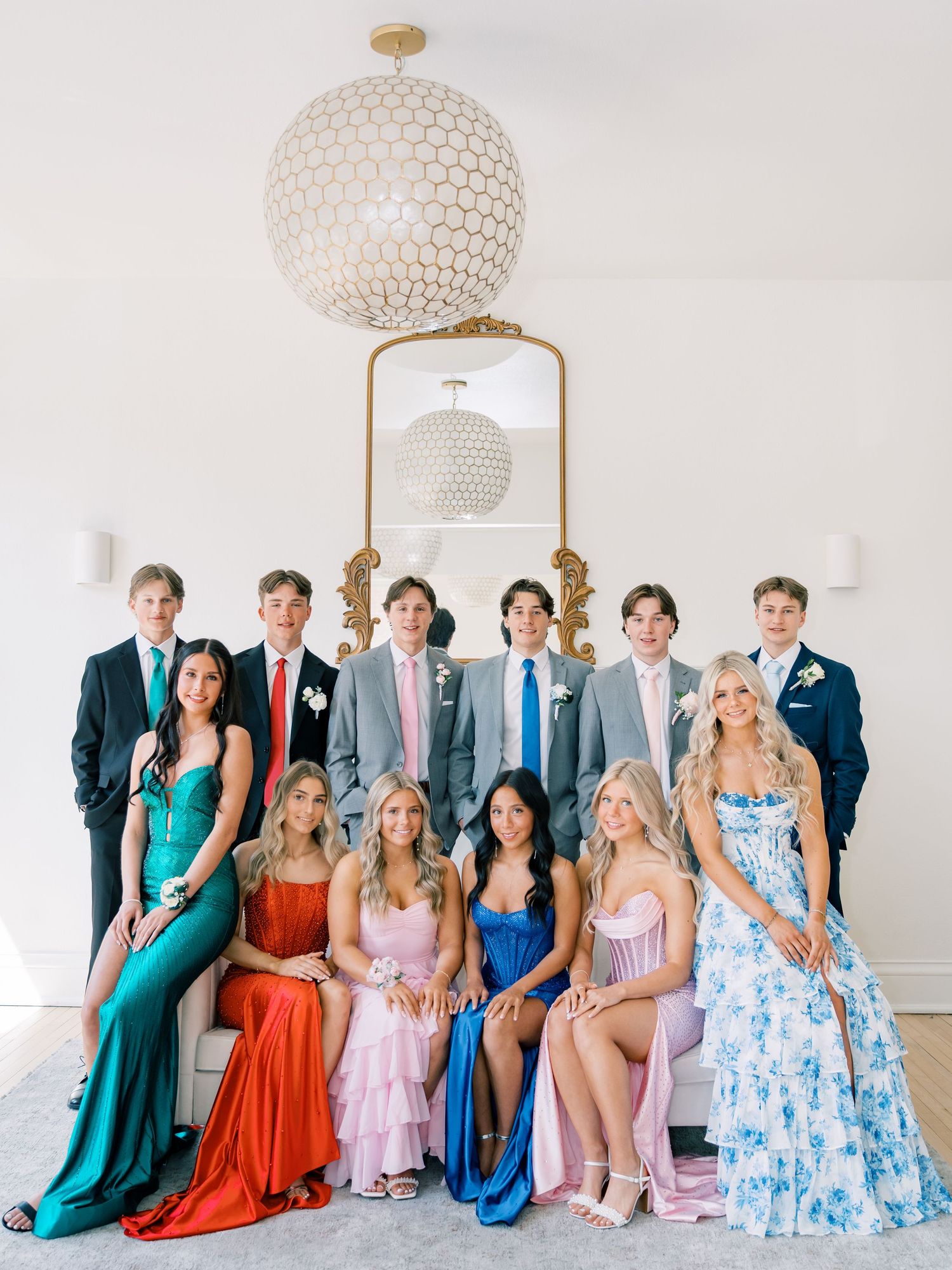Group of teens in formal prom attire posing together, girls in colorful gowns seated, boys in suits standing behind.