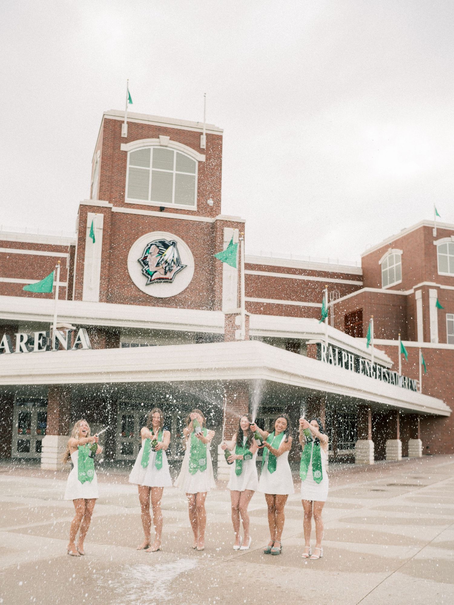 Six women in white dresses with green sashes celebrate outside Ralph Engelstad Arena, popping champagne in the snow.