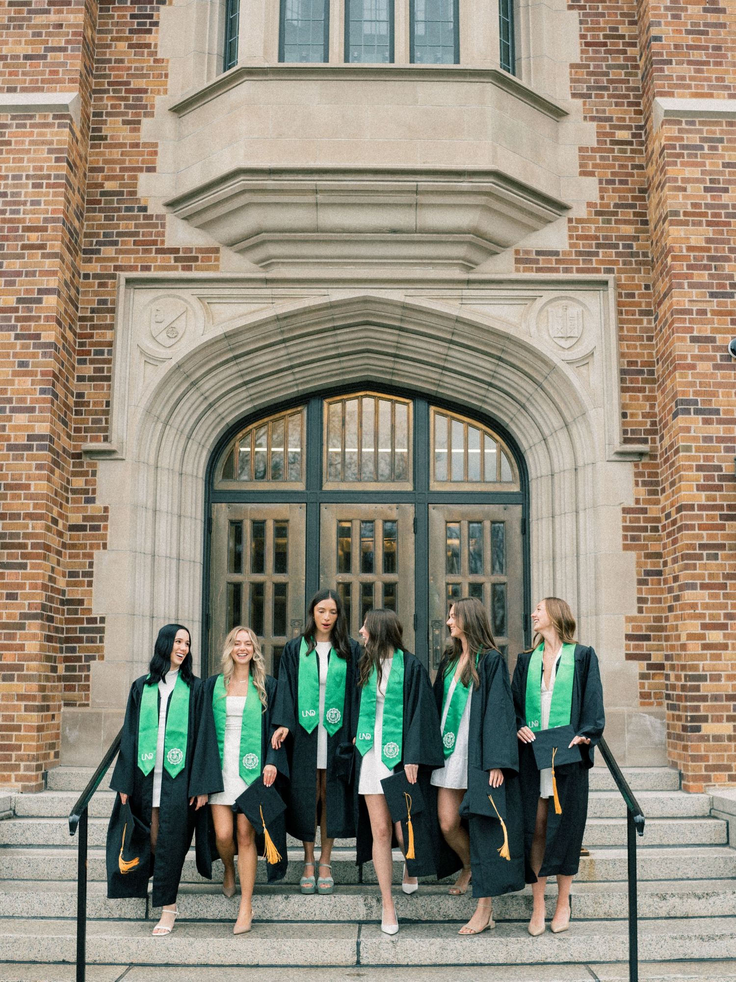 Six female graduates in black and green caps and gowns pose on steps of a brick university building.