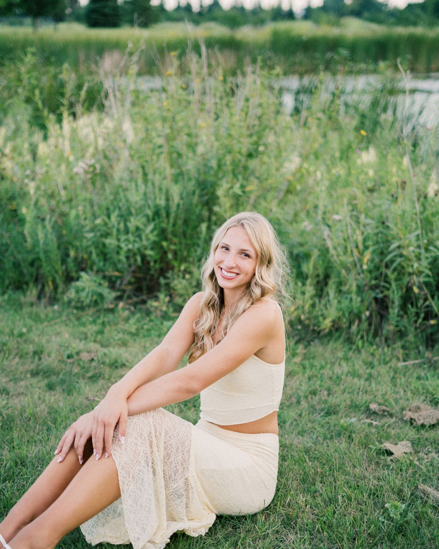 A person in a cream-colored dress sits gracefully on grassy ground during an outdoor photoshoot.