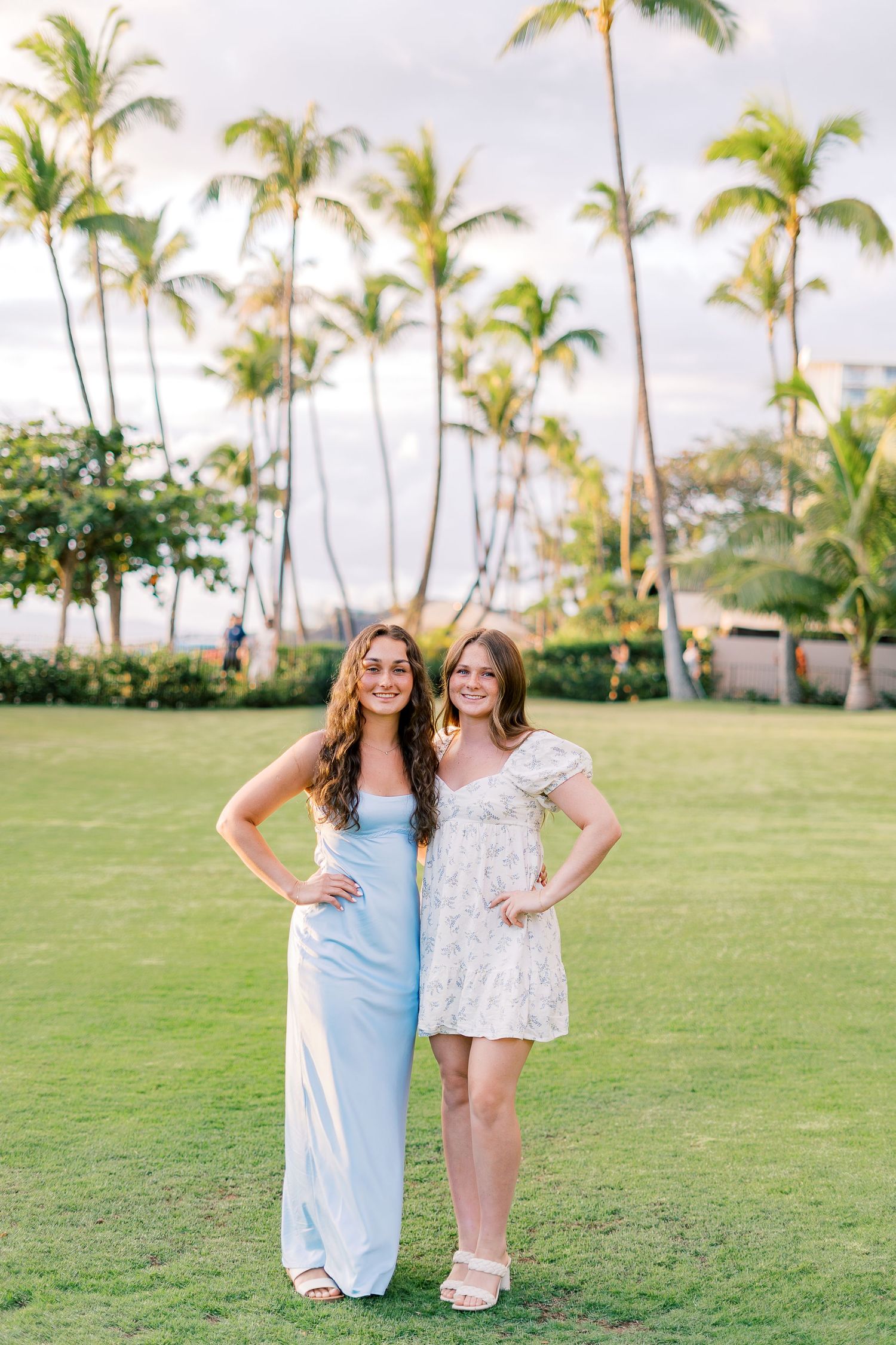 Two friends pose together on a lush lawn with tropical palm trees in Hawaii.