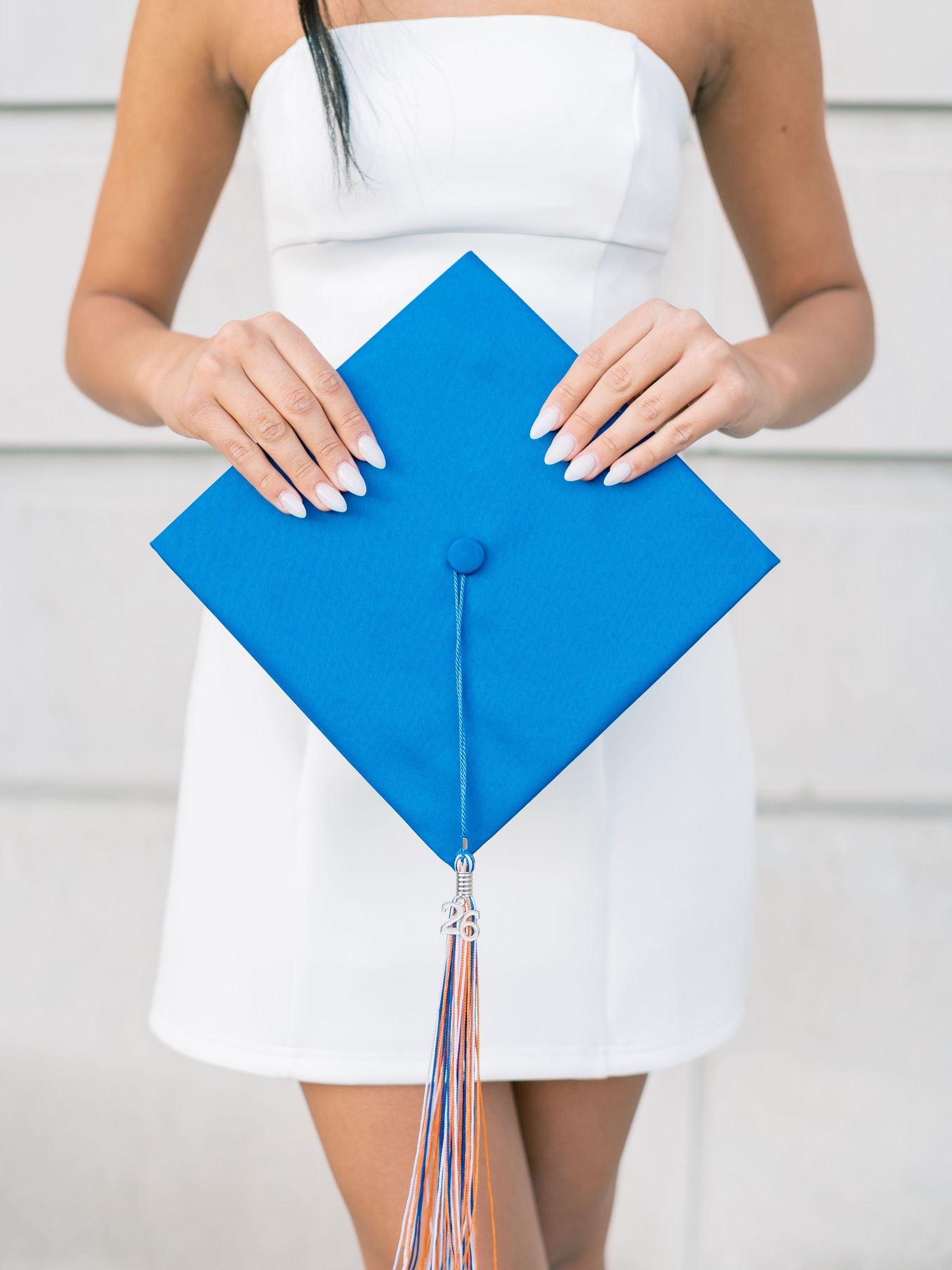Woman in white dress holding a blue graduation mortarboard cap with colorful tassel.