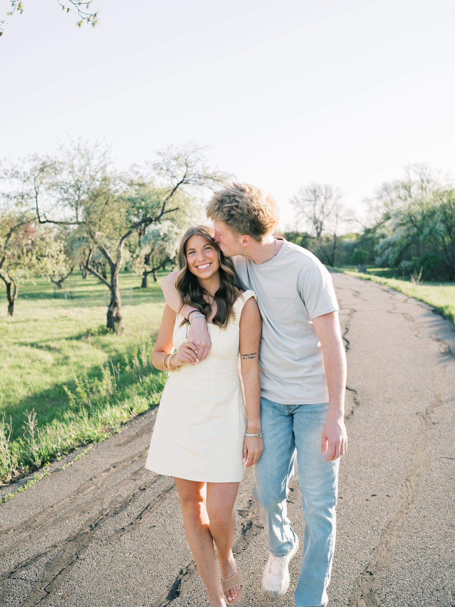 A couple walks hand-in-hand down a sunny dirt road, sharing a kiss, surrounded by lush green trees.