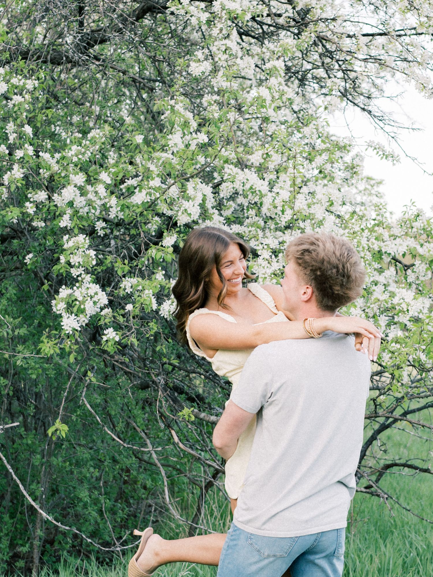A joyful couple embraces near a blooming white flowering tree in a lush green outdoor setting.