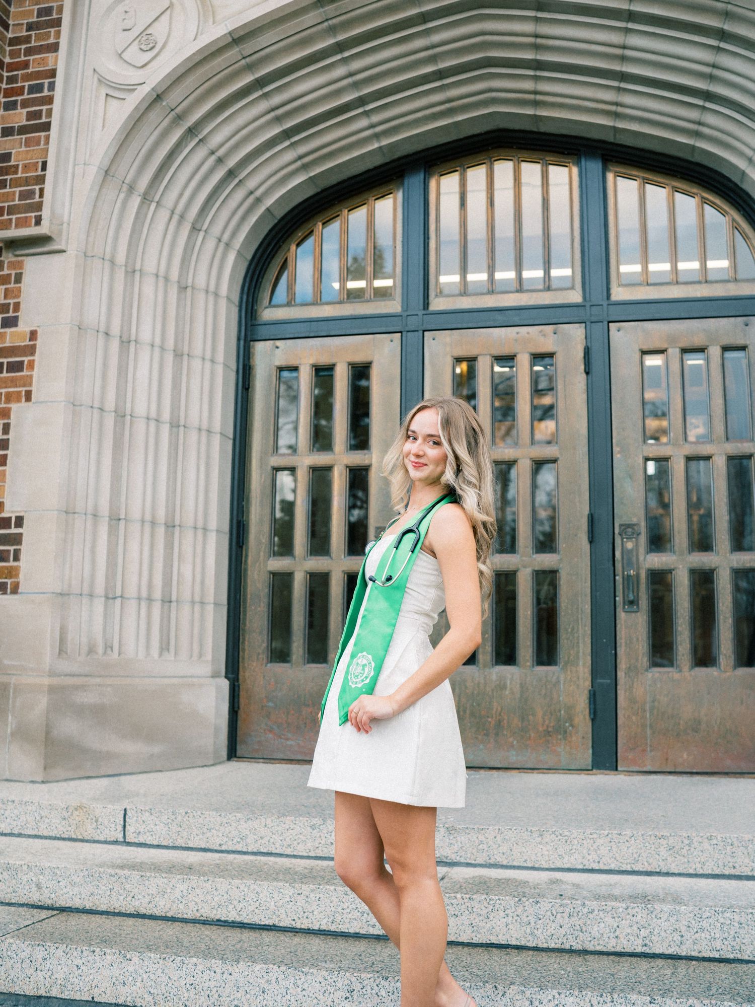 Smiling graduate in white dress and green graduation stole poses on steps before ornate arched building entrance.