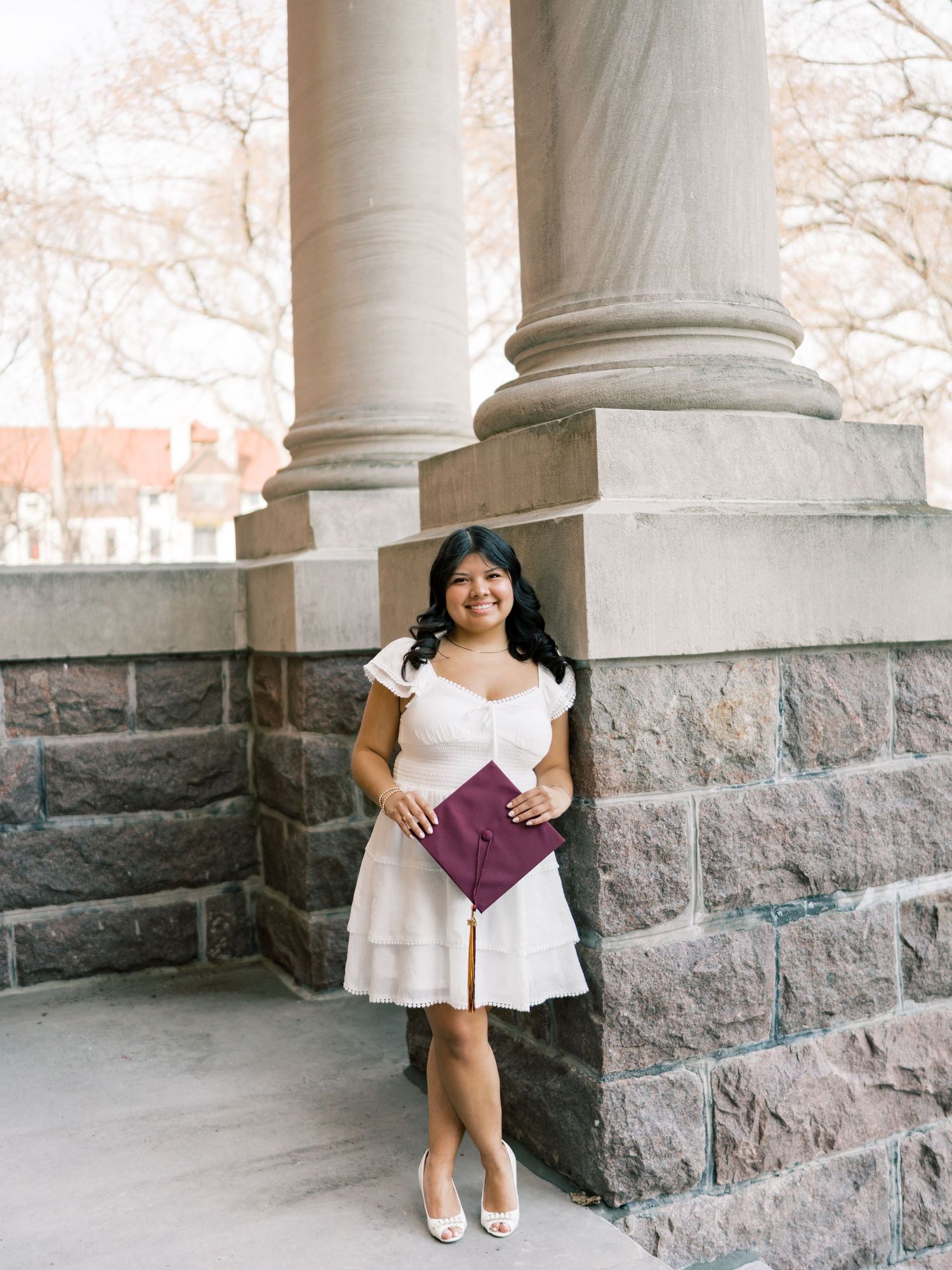 Young woman in white dress holding burgundy graduation cap poses between stone columns at university building.