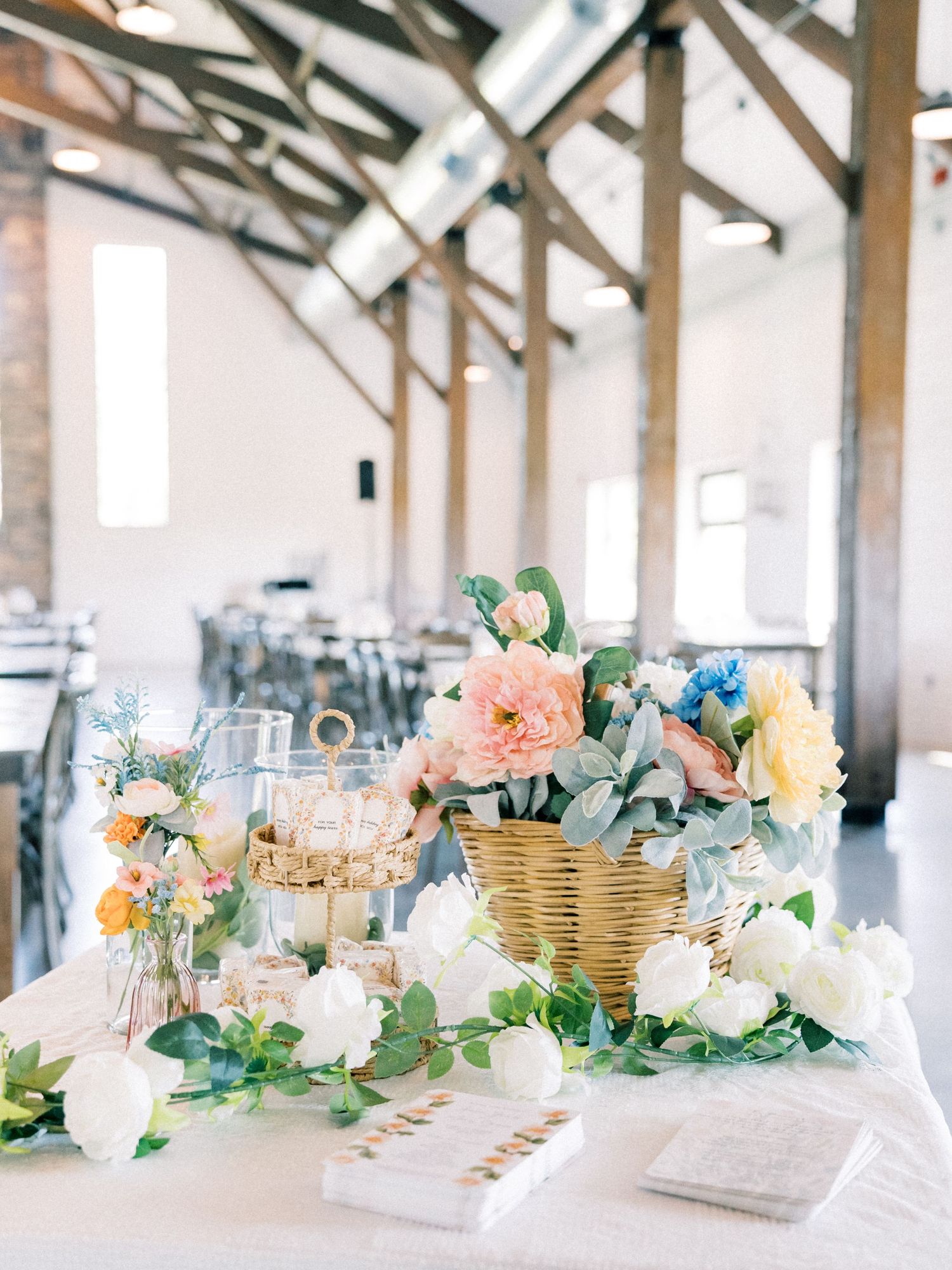 Elegant wedding reception table with colorful floral arrangements in a wicker basket, white roses, and greenery in a bright barn venue.