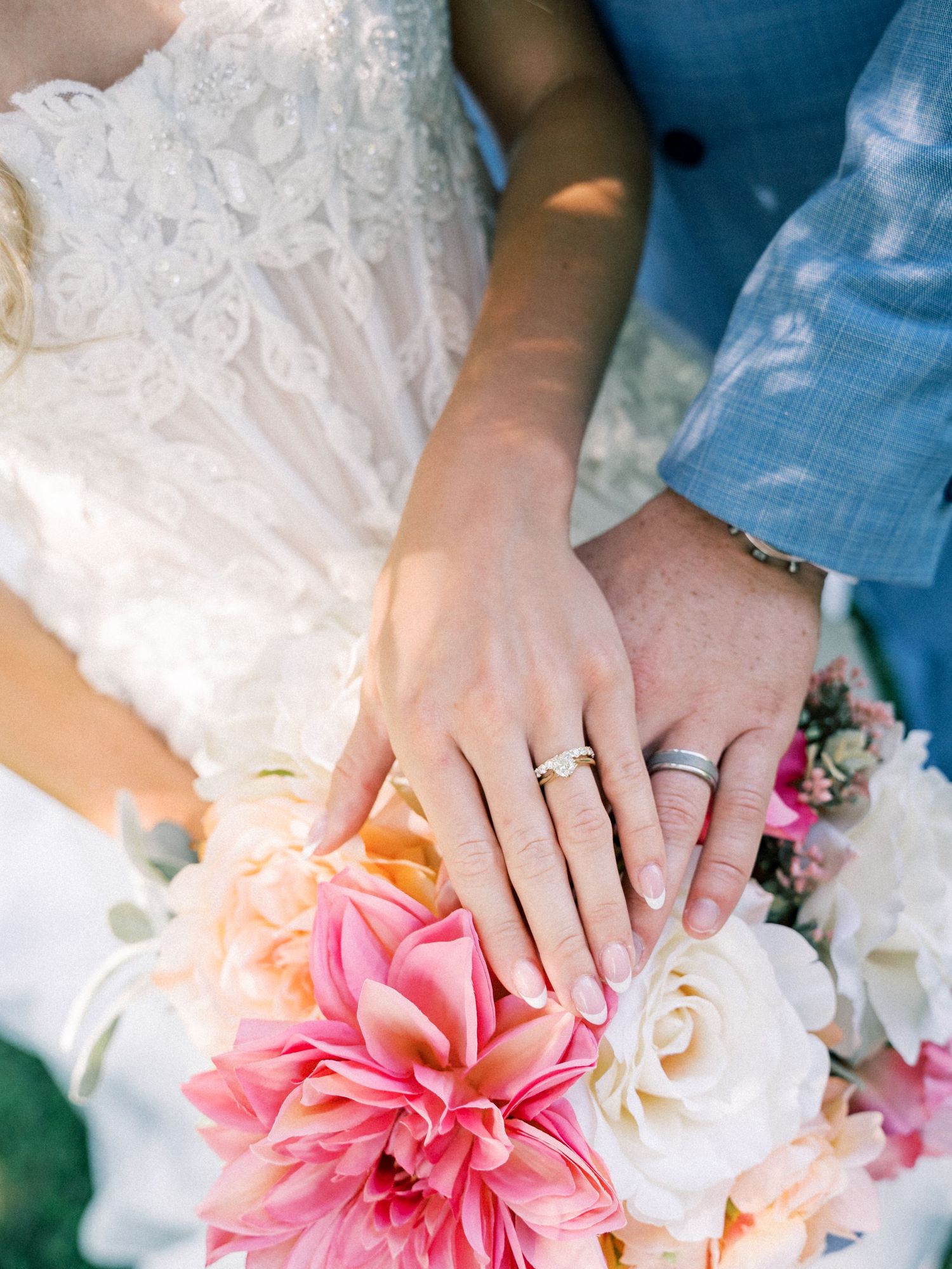 Bride and groom hands with wedding rings resting on a colorful bridal bouquet of pink dahlias and white roses.