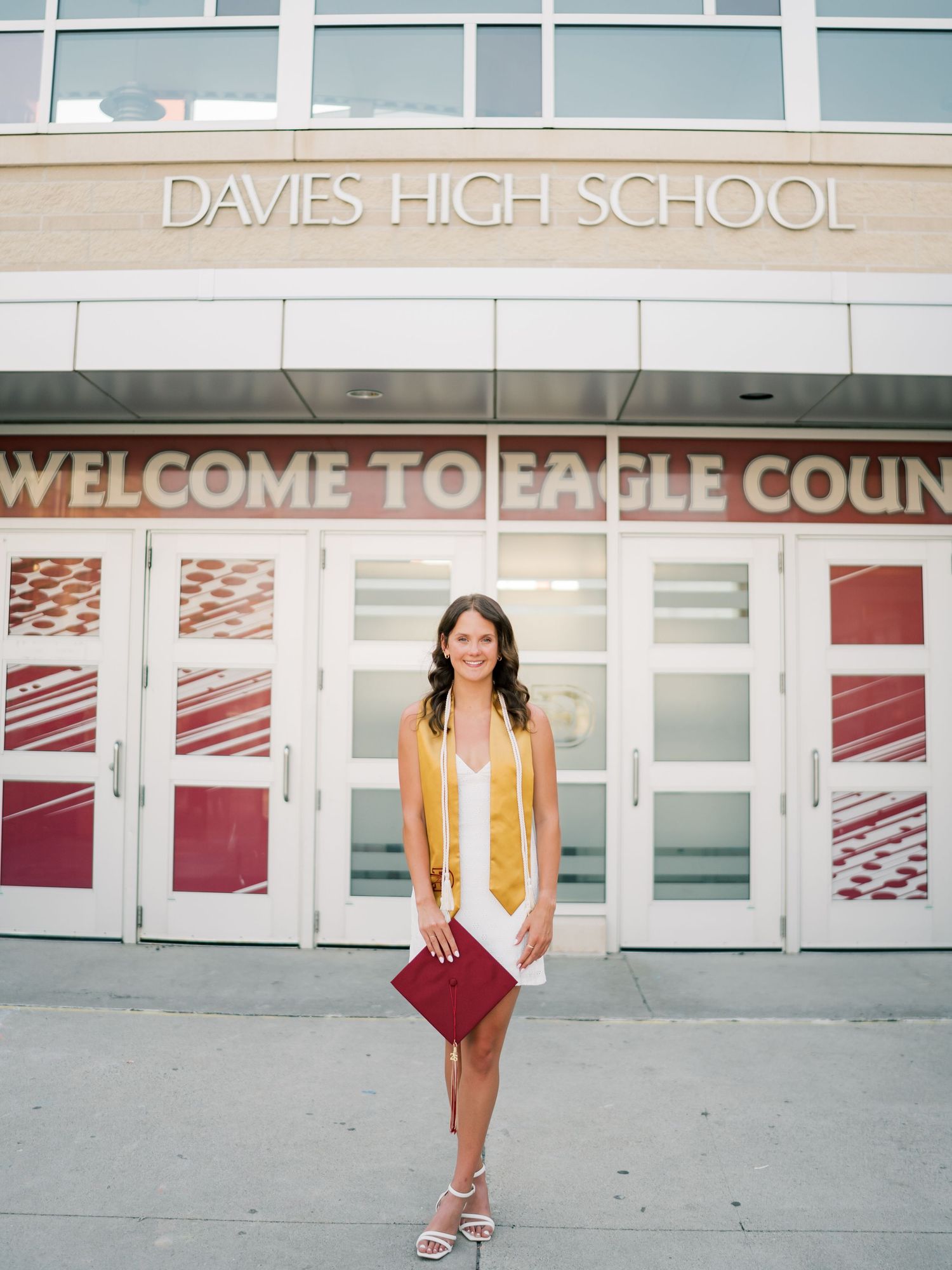 High school graduate poses in white dress and gold honor stole holding maroon cap outside Davies High School entrance.