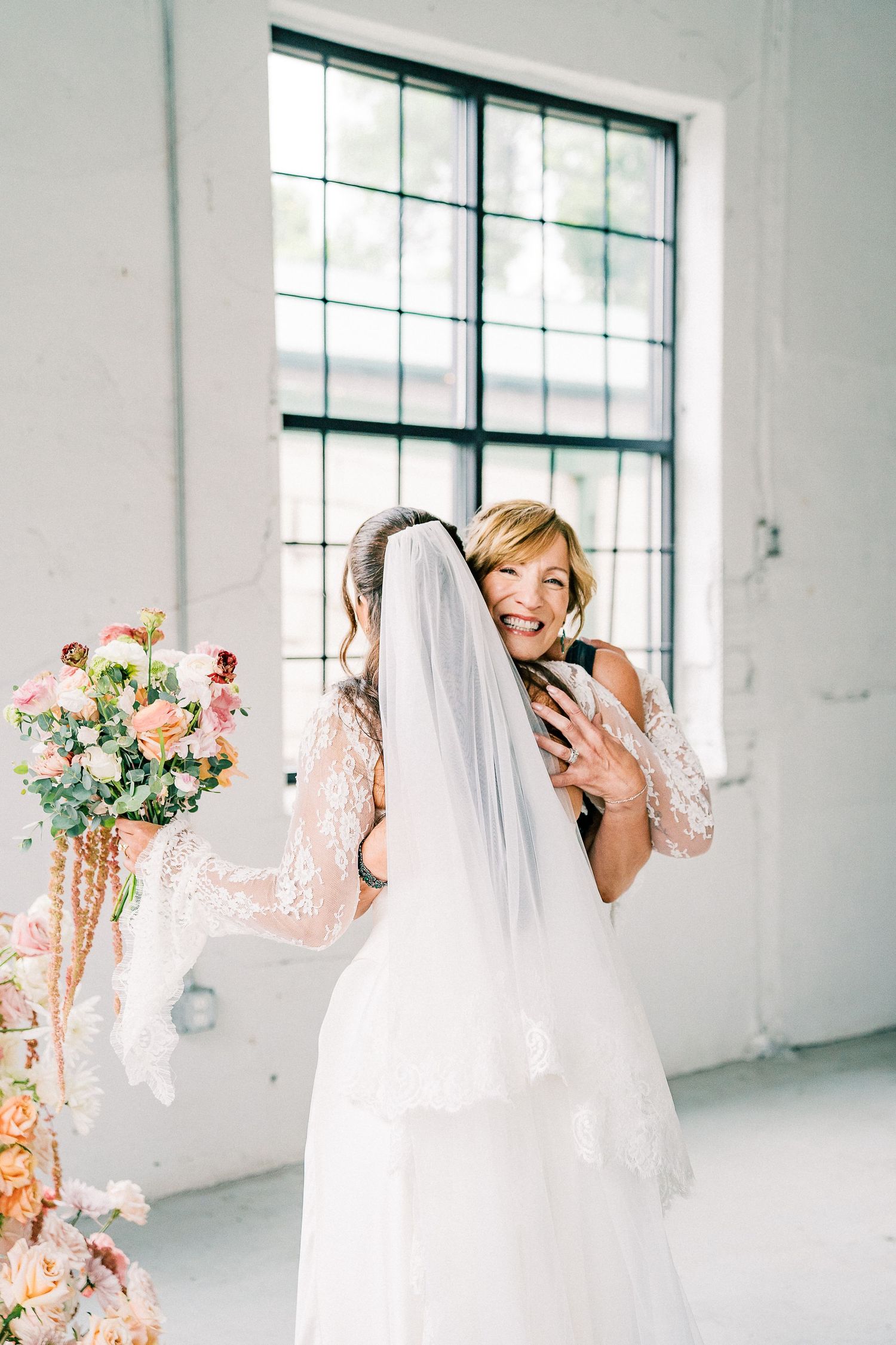 A joyful bride in a white lace gown and veil laughs candidly in a bright, minimalist studio space.