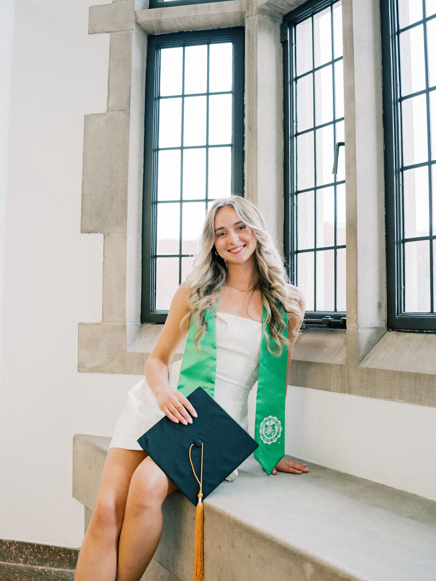 Smiling graduate in white dress with green sash holds cap on stone steps near tall windows.
