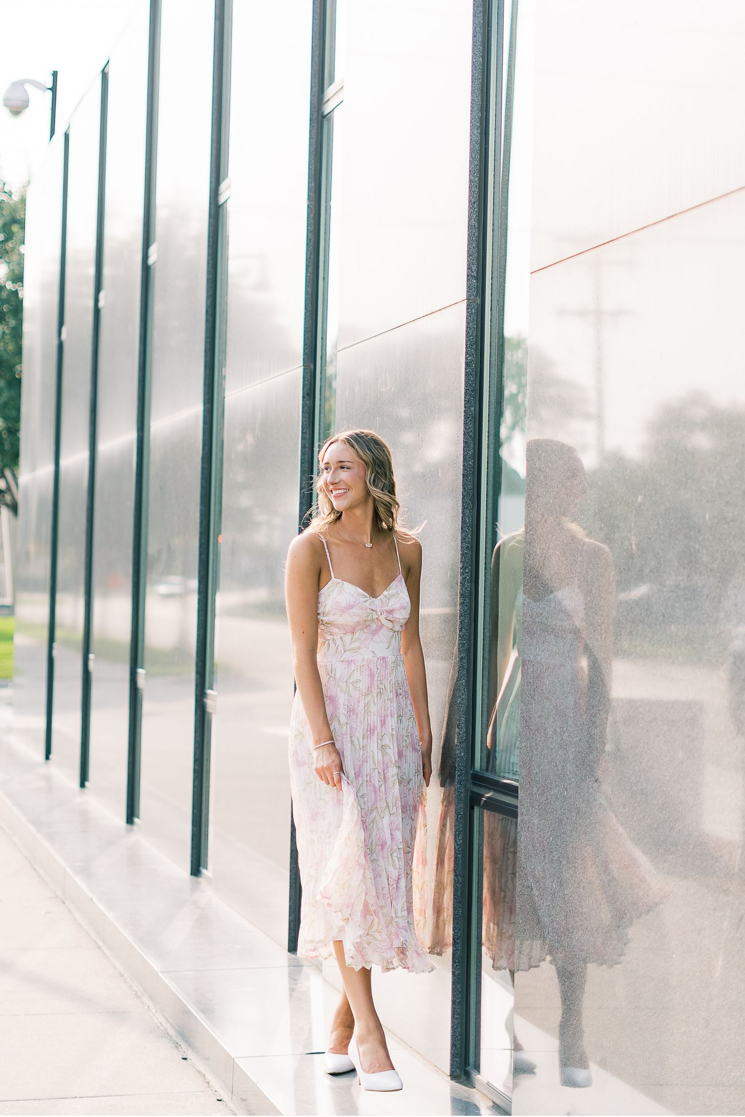 Woman in a white floral sundress poses against a modern glass and metal building exterior in a stylish photoshoot.