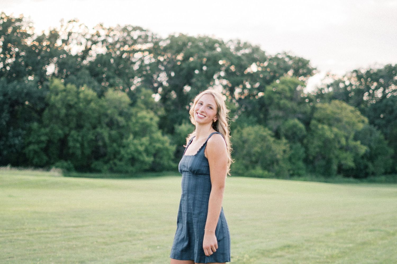 A person in a navy blue dress stands in a lush green field during golden hour lighting.